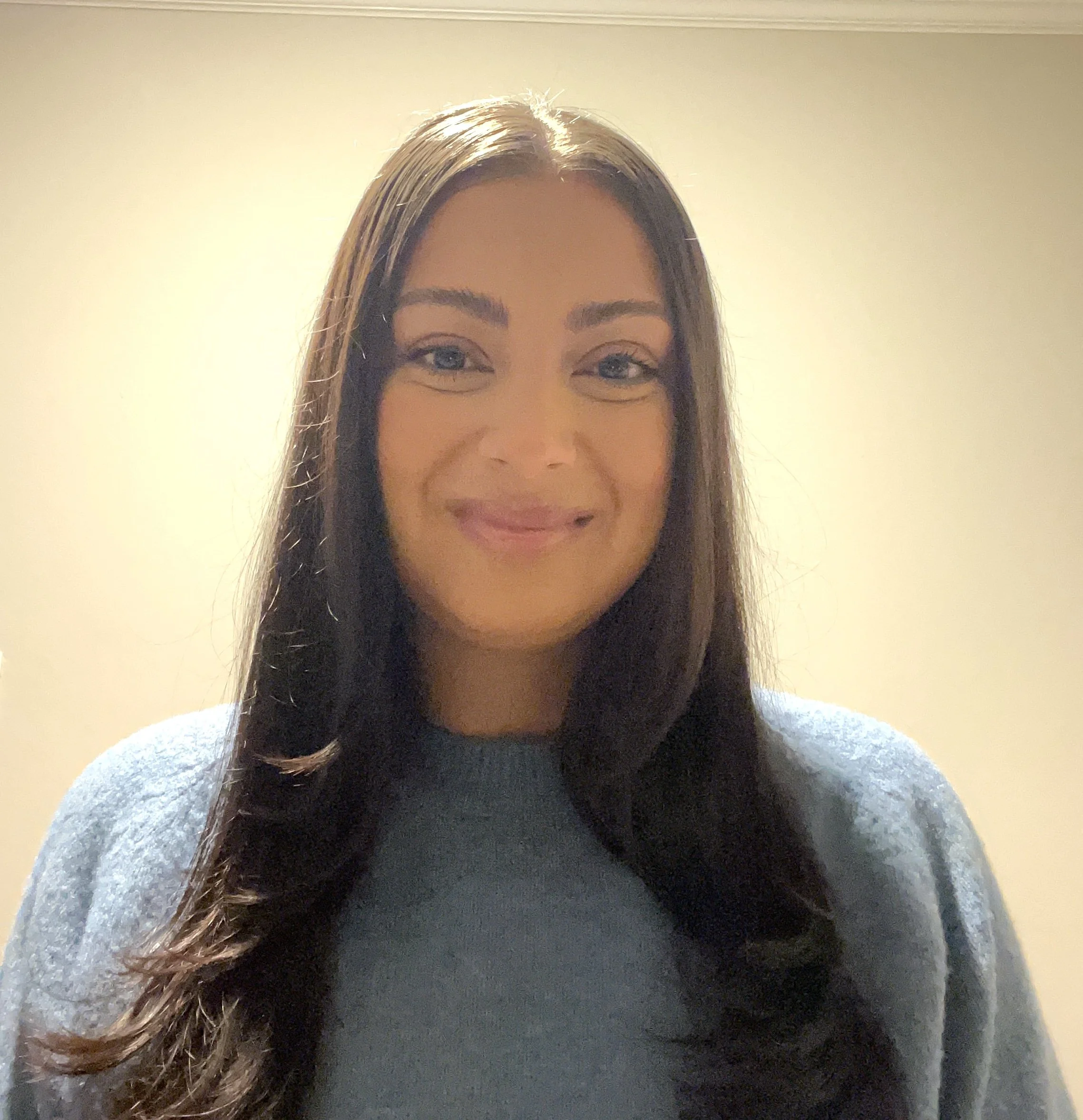 Close-up of a woman's upper face, smiling, with long brown hair, blue eyes, and light makeup, standing against a plain light-colored wall.