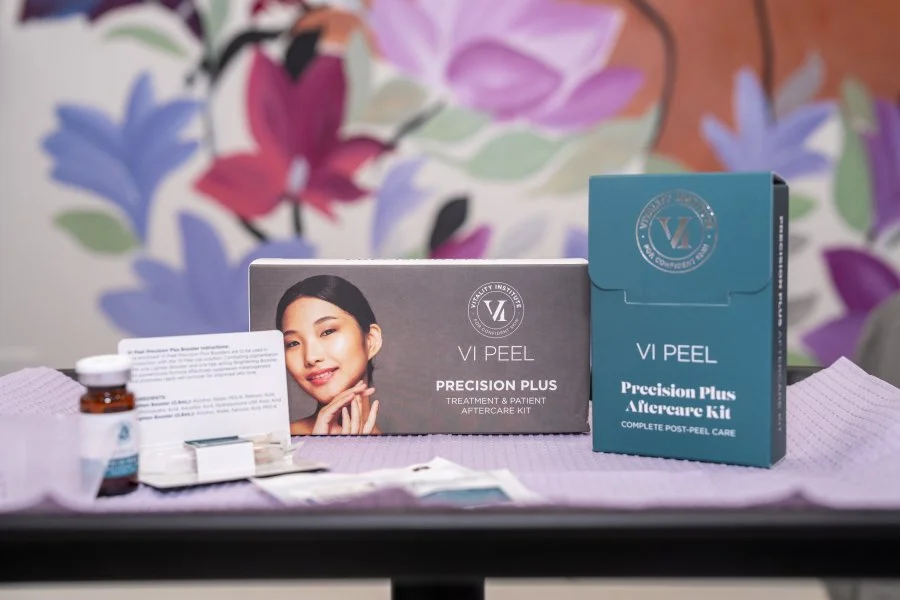 Boxes of VI Peel skincare products, including a Precision Plus Treatment & Patient Aftercare Kit, on a table with medical supplies and a colorful floral mural in the background.