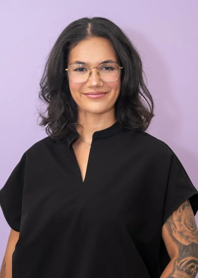A woman with shoulder-length dark hair, glasses, wearing a black top, smiling against a light purple background.