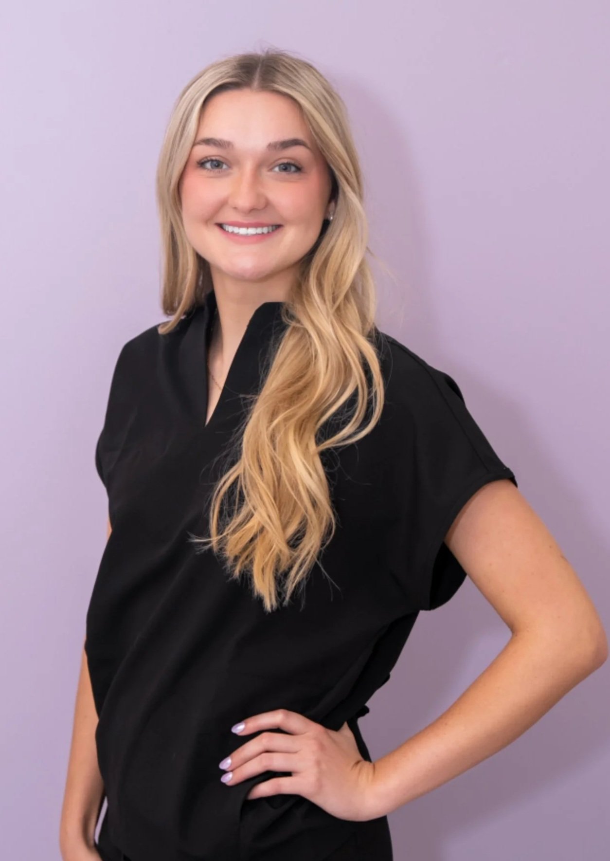 A smiling young woman with long wavy blonde hair, wearing a black shirt, standing against a light purple background.