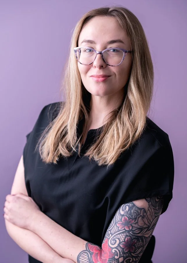 A woman with long blonde hair, wearing glasses and a black top, standing against a purple background, with her arms crossed, showing a colorful floral tattoo on her left arm.
