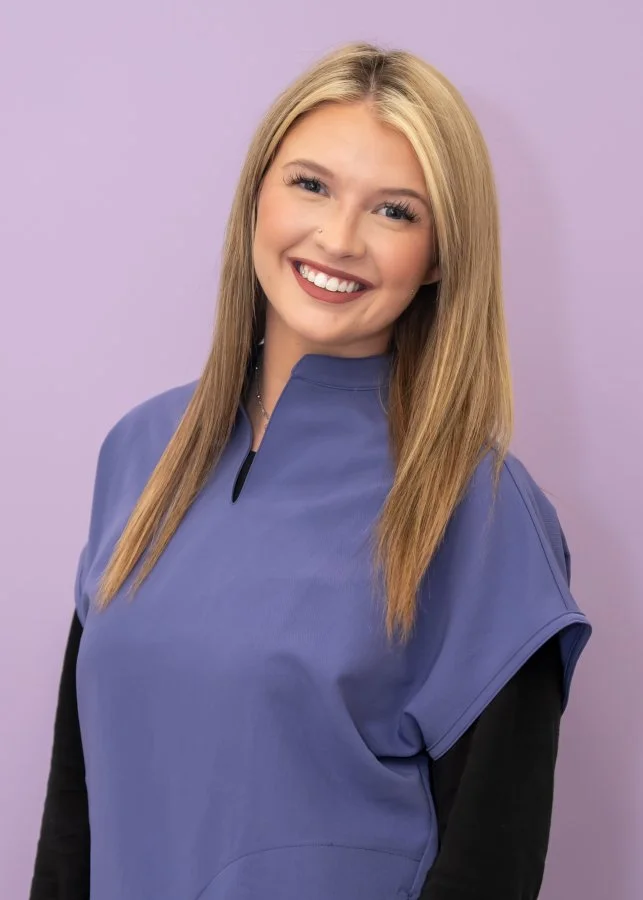A young woman with long blonde hair, smiling, wearing a blue short-sleeved top over a black long-sleeved shirt, standing against a light purple background.