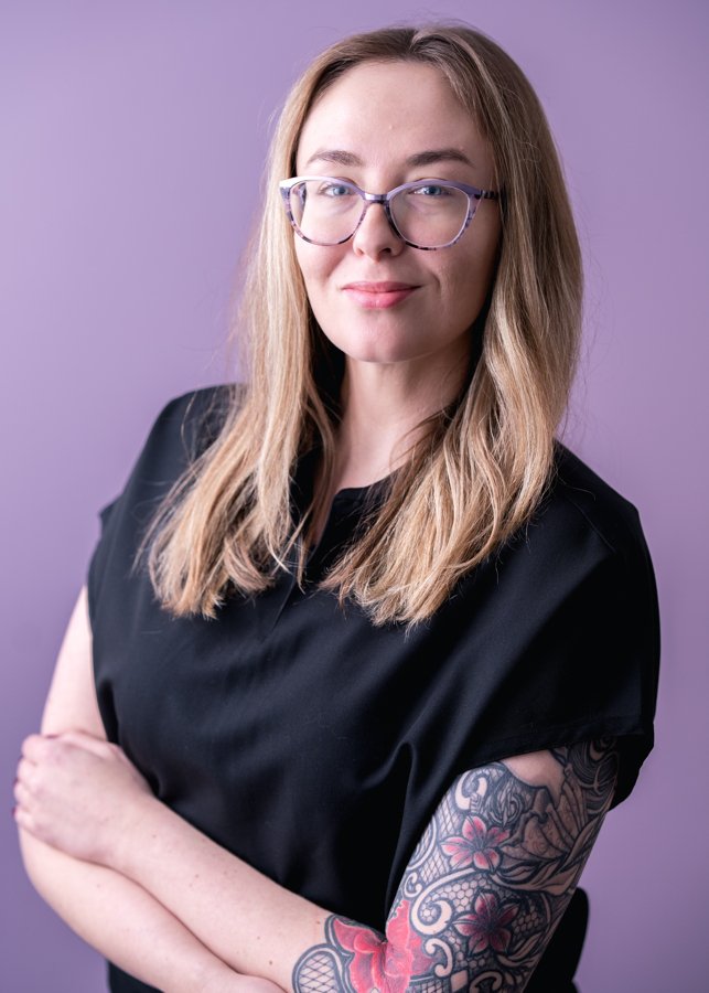 A young woman with long blonde hair, glasses, and a tattooed arm, wearing a black top, standing with arms crossed against a purple background.
