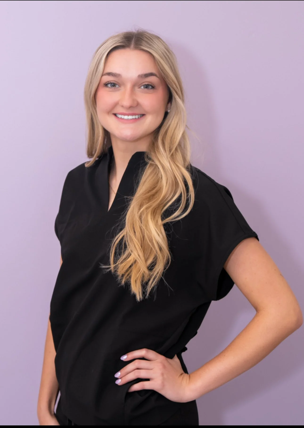 Portrait of a young woman with long blonde hair, smiling, wearing a black short-sleeved top and standing against a light purple background.