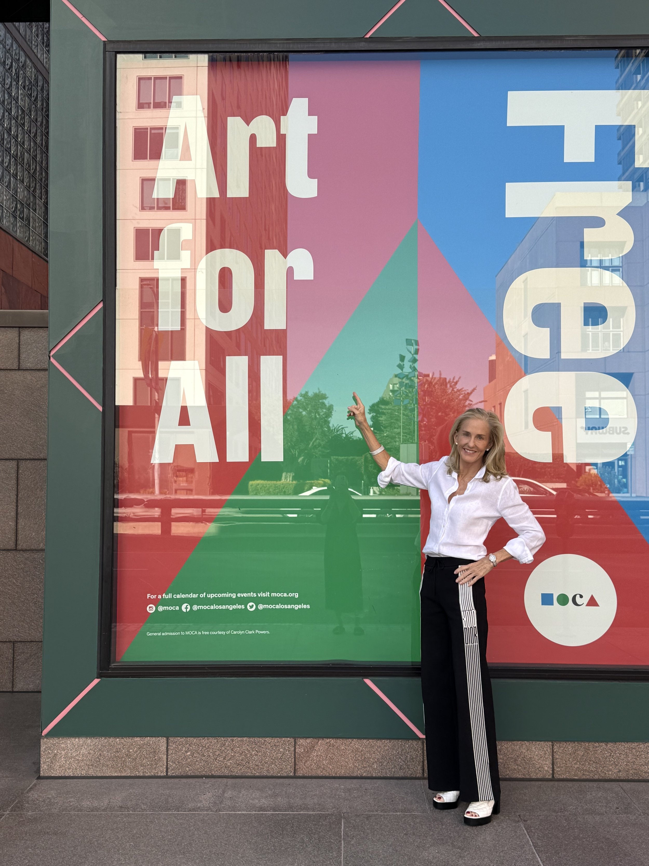 A woman standing in front of a colorful art display window that reads 'Art for All' and 'MOCA' with her right arm raised and pointing at the sign.