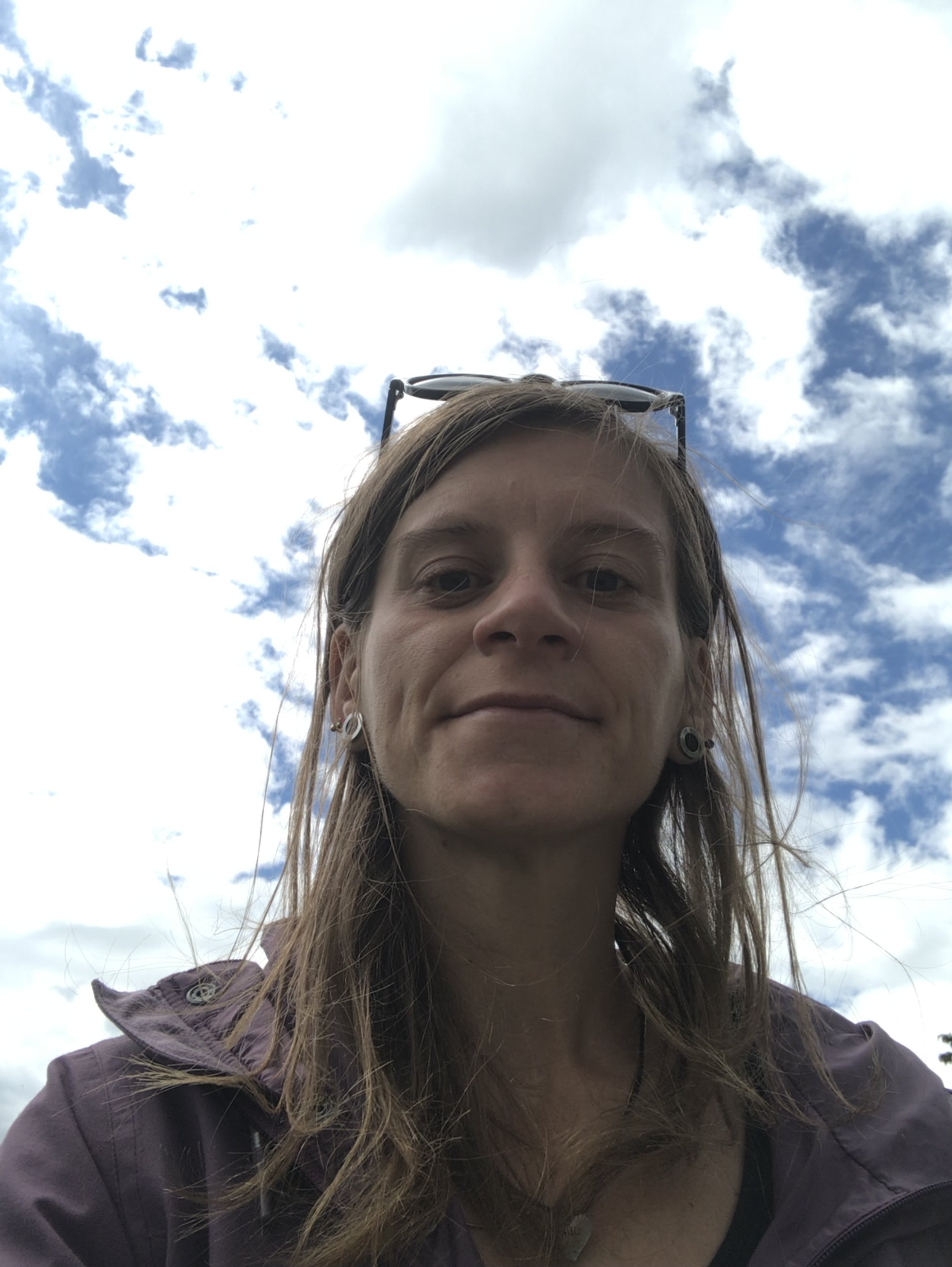 Close-up of a woman outdoors, looking down at the camera, with a partly cloudy sky in the background. She has long hair, earrings, and sunglasses on her head.
