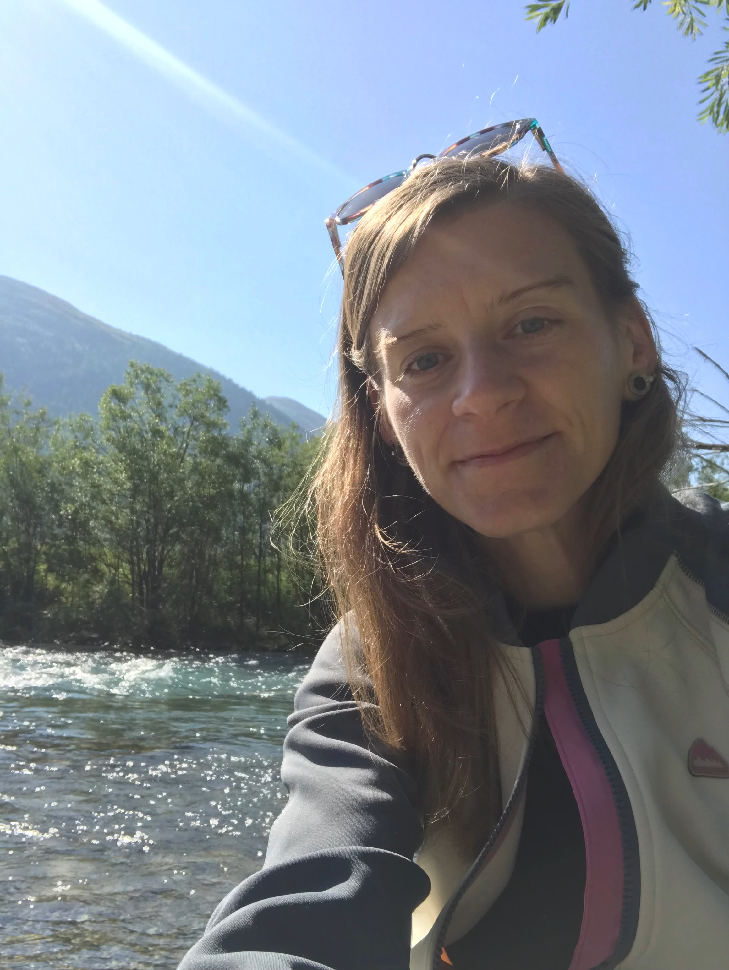 A woman with long brown hair, wearing sunglasses on her head and a jacket, takes a selfie outdoors near a river with trees and mountains in the background on a sunny day.
