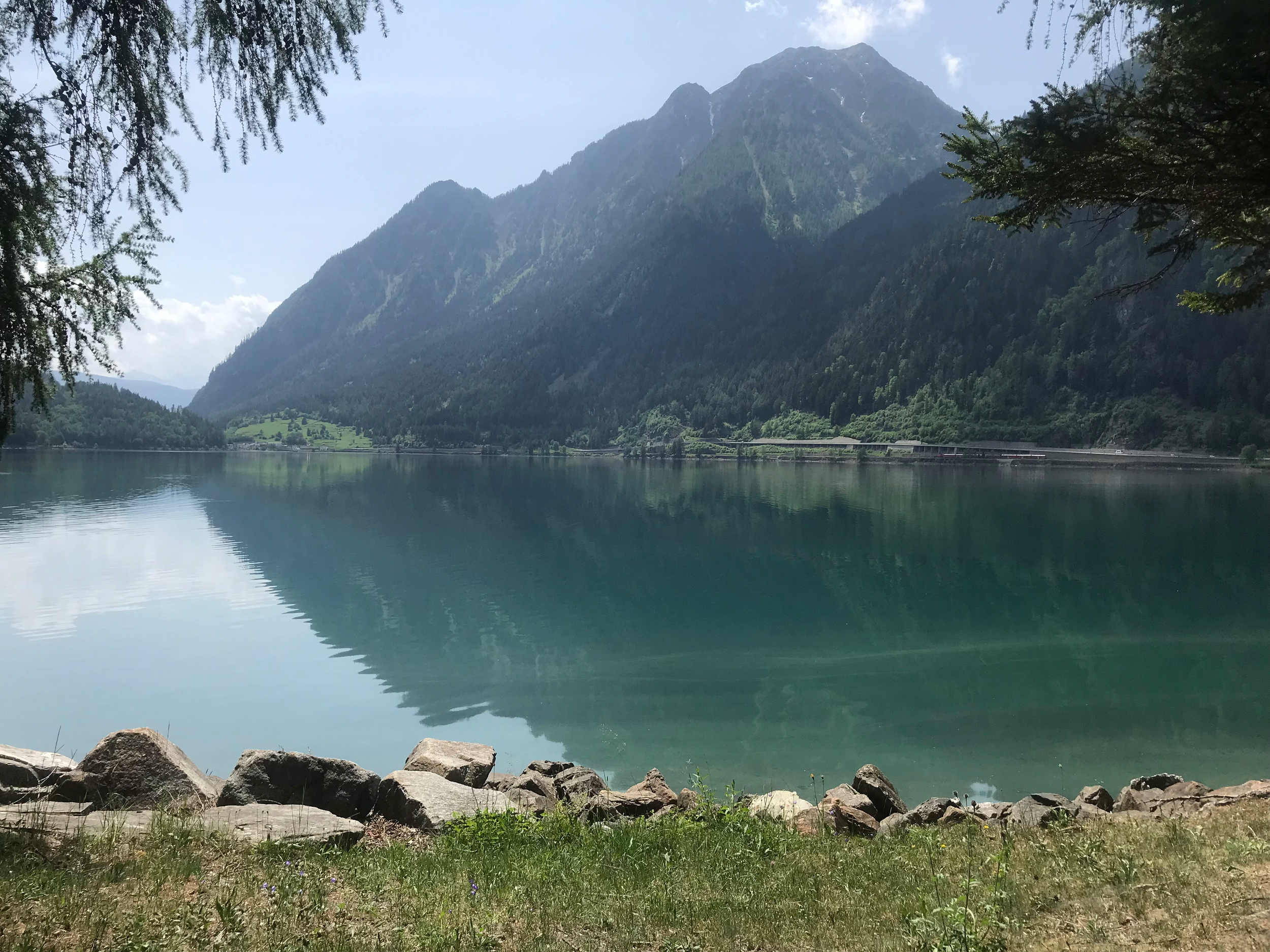 A tranquil mountain lake with clear blue water, surrounded by rocky shoreline, green grass, and overhanging trees, with tall mountains covered in dense forest in the background under a partly cloudy sky.