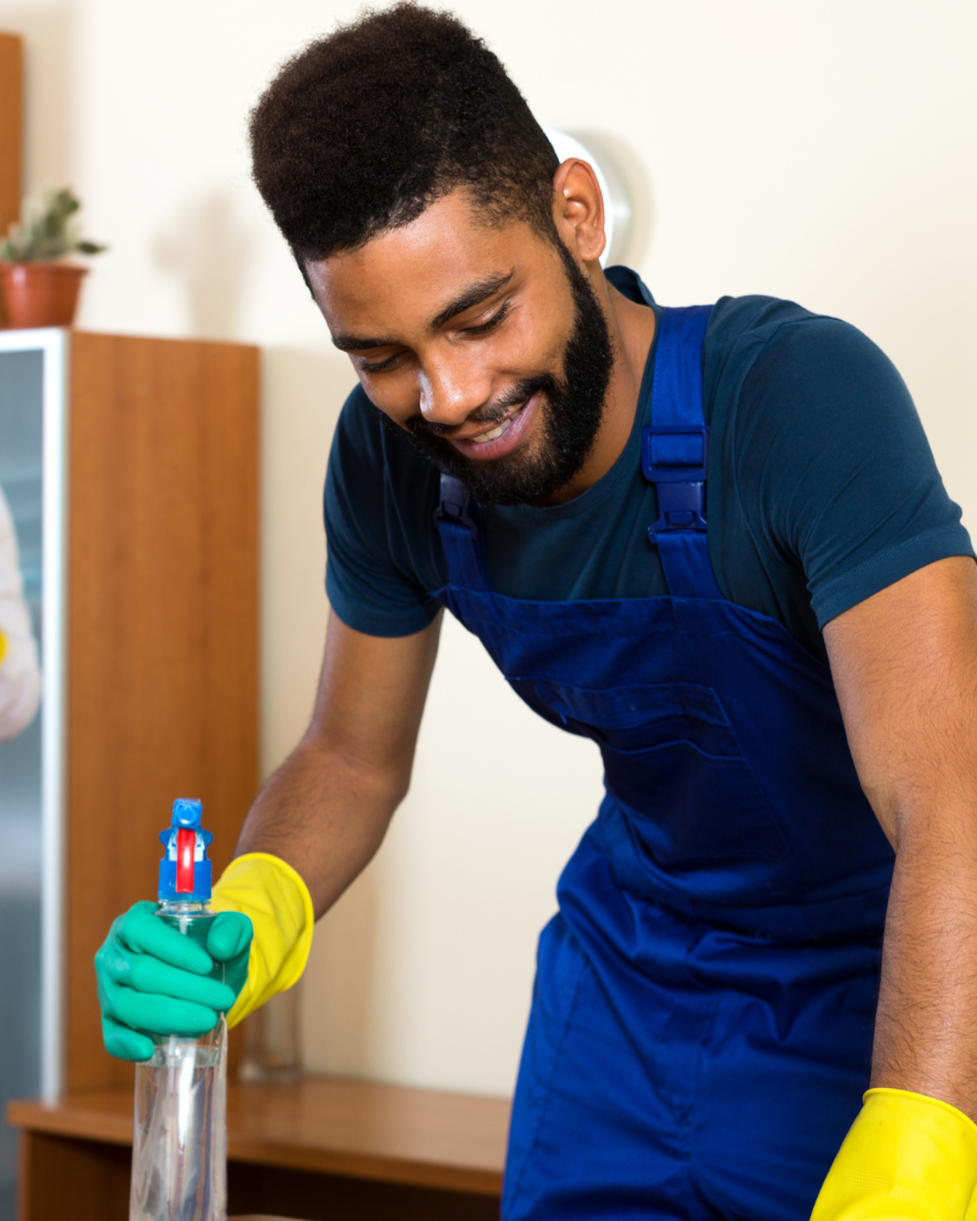A man wearing a navy blue shirt, blue overalls, and yellow and green rubber gloves is smiling while working with a glass container. He appears to be engaged in a cleaning or scientific task in a room with a wooden cabinet and a potted plant in the background.