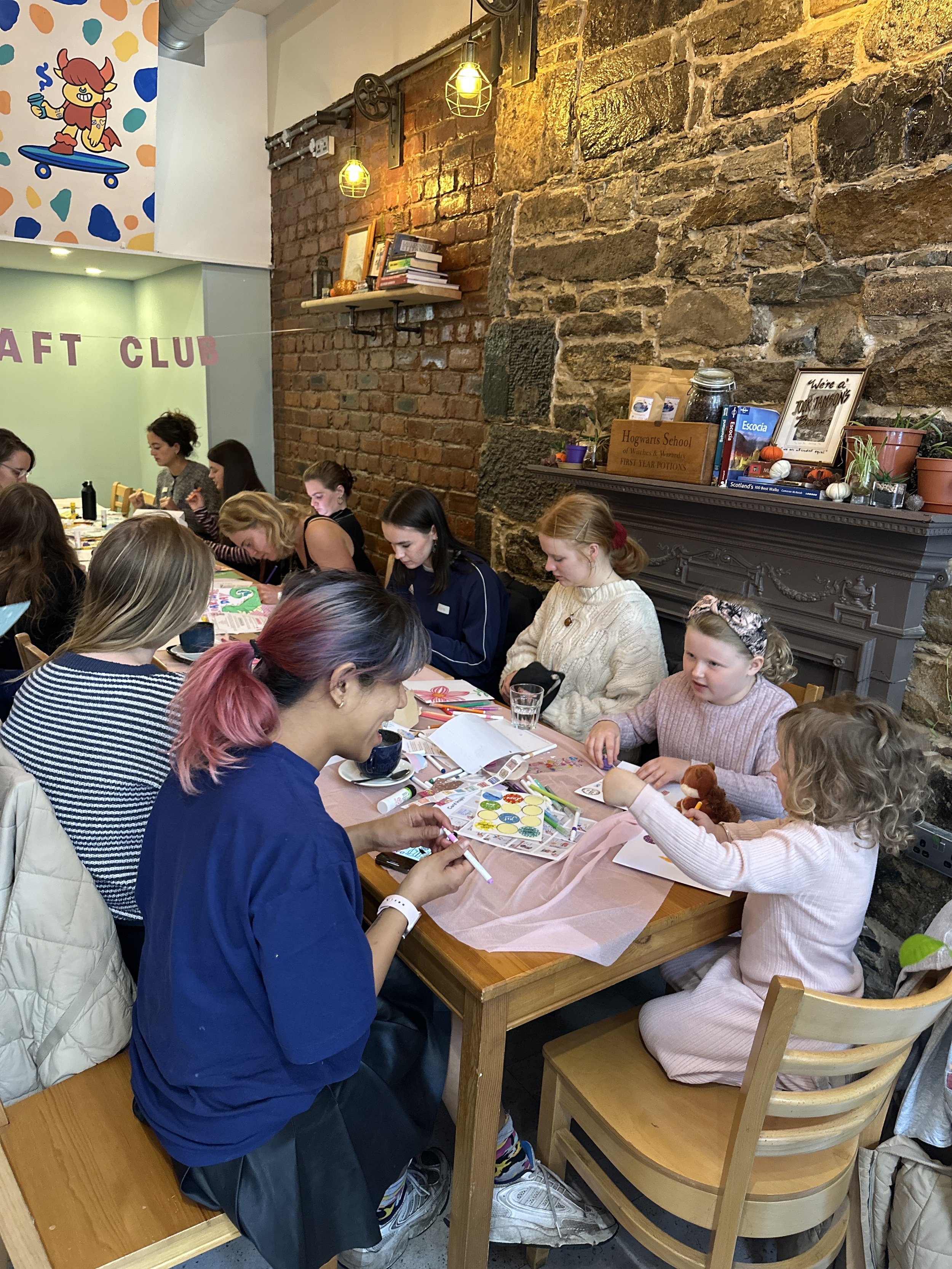 Children and adults sitting at a table engaged in arts and crafts activities inside a cozy room with brick walls, decorative shelves, and warm lighting.