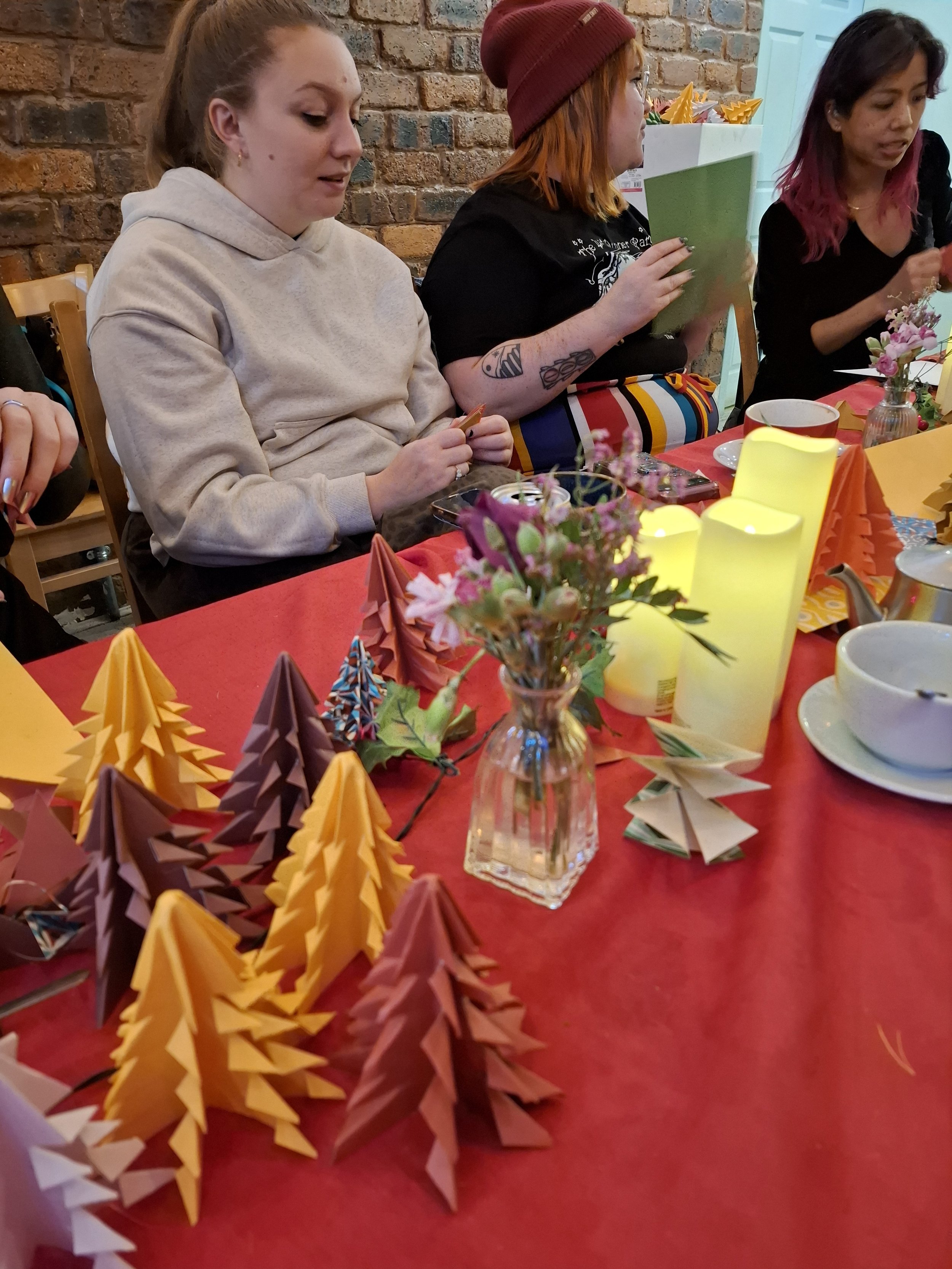 Three women seated at a holiday table with origami paper Christmas trees, candles, a vase of flowers, and tableware, with a brick wall and a teal door in the background.