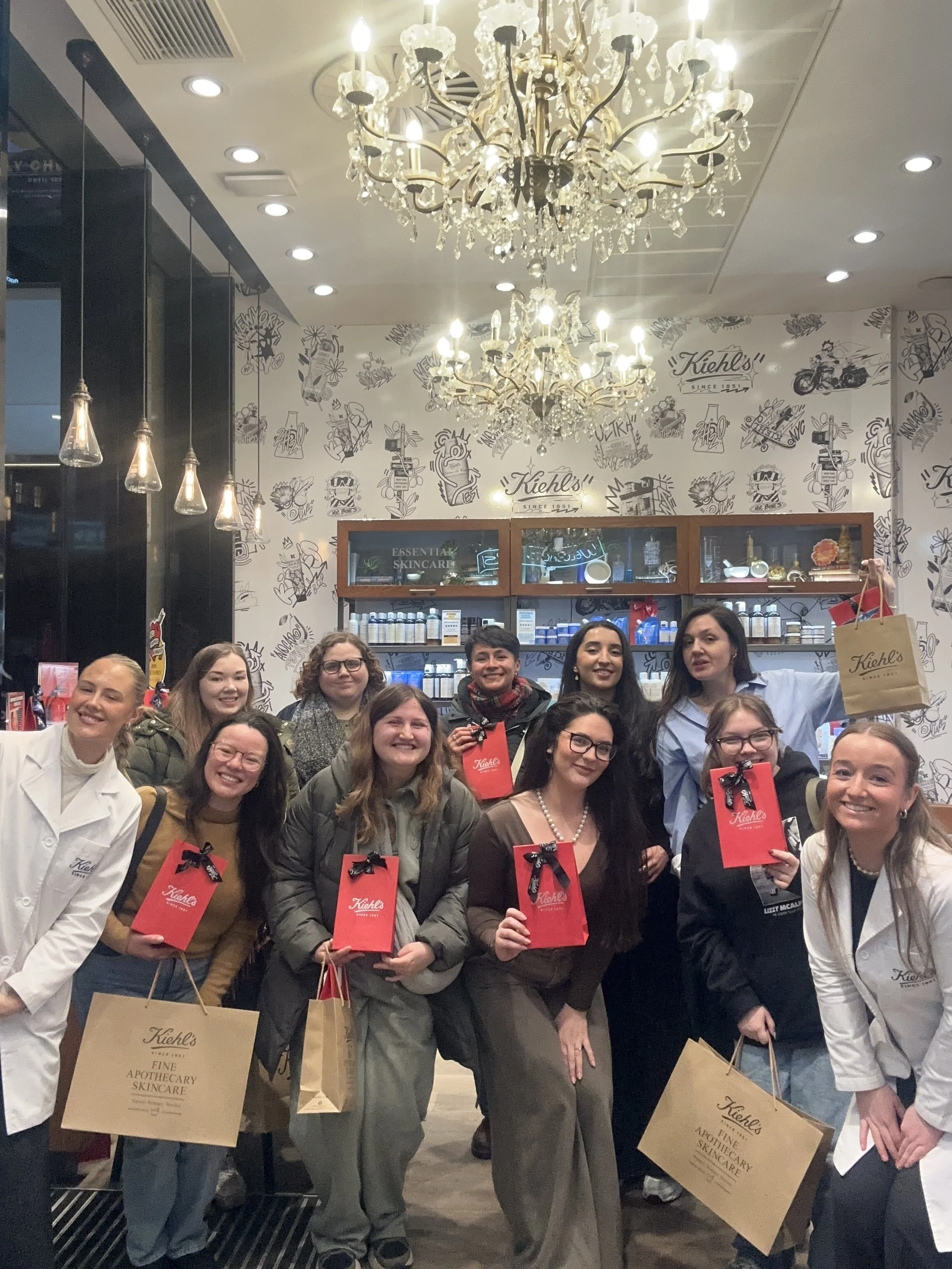 Three women seated at a holiday table with origami paper Christmas trees, candles, a vase of flowers, and tableware, with a brick wall and a teal door in the background.