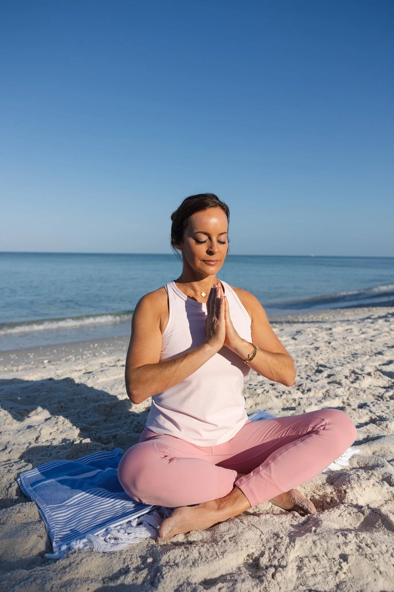 Woman practicing yoga on a beach, sitting cross-legged with hands in prayer position, eyes closed, wearing a white tank top and pink pants.