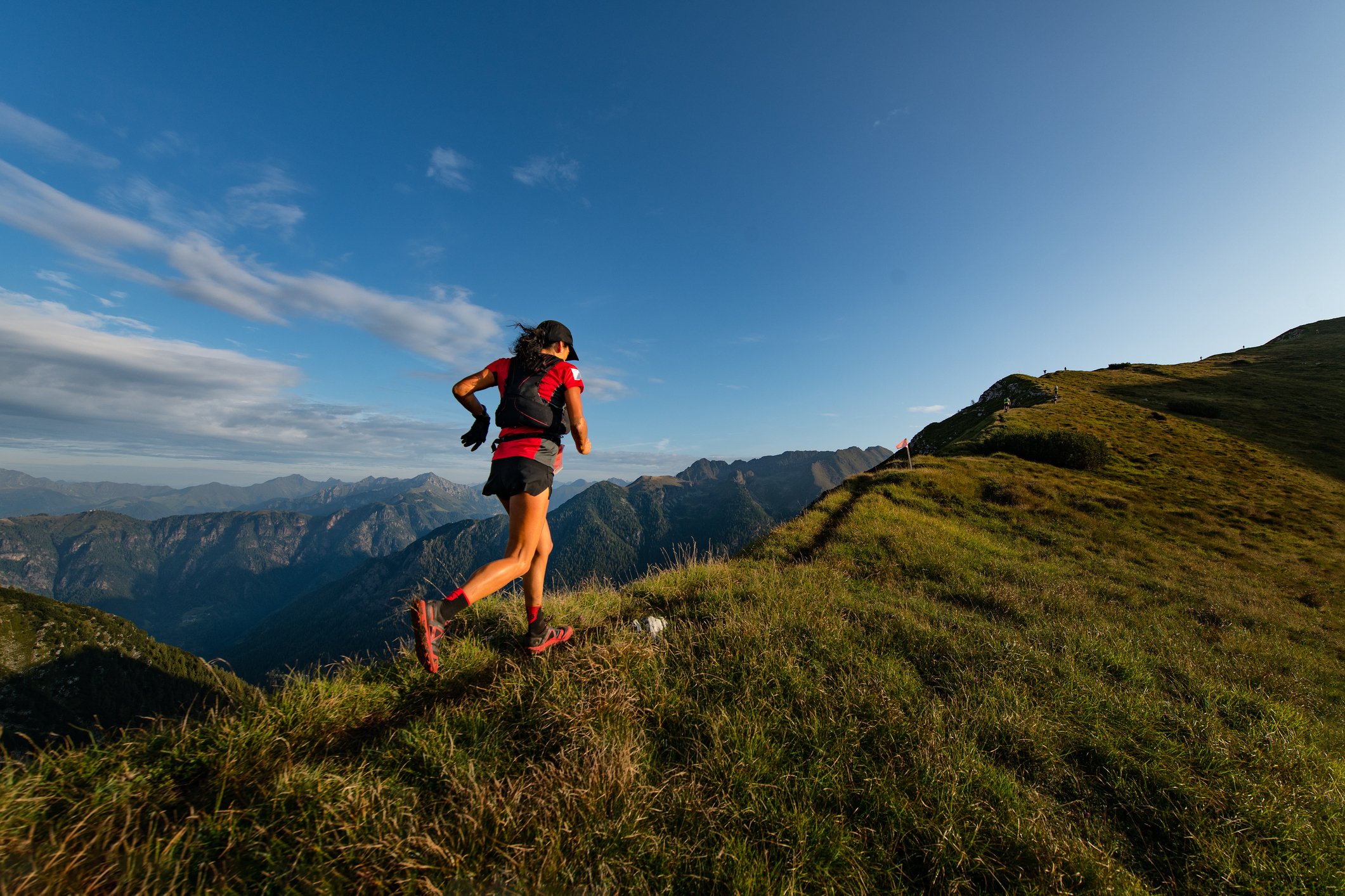 A female trail runner wearing a red shirt, black shorts, and a backpack running along a grassy mountain trail with a mountain range in the background on a clear day. Shown in relation to a sports nutritionist working with endurance athletes.