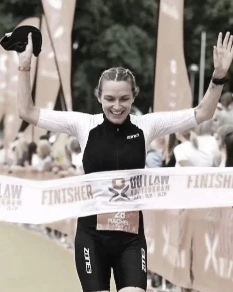 A  triathlete crossing the finish line of Outlaw Triathlon with her arms raised in victory, smiling, with a banner reading 'FINISHER' in front of her. Related to sports nutrition and triathlon nutrition advice from a registered sports nutritionist.