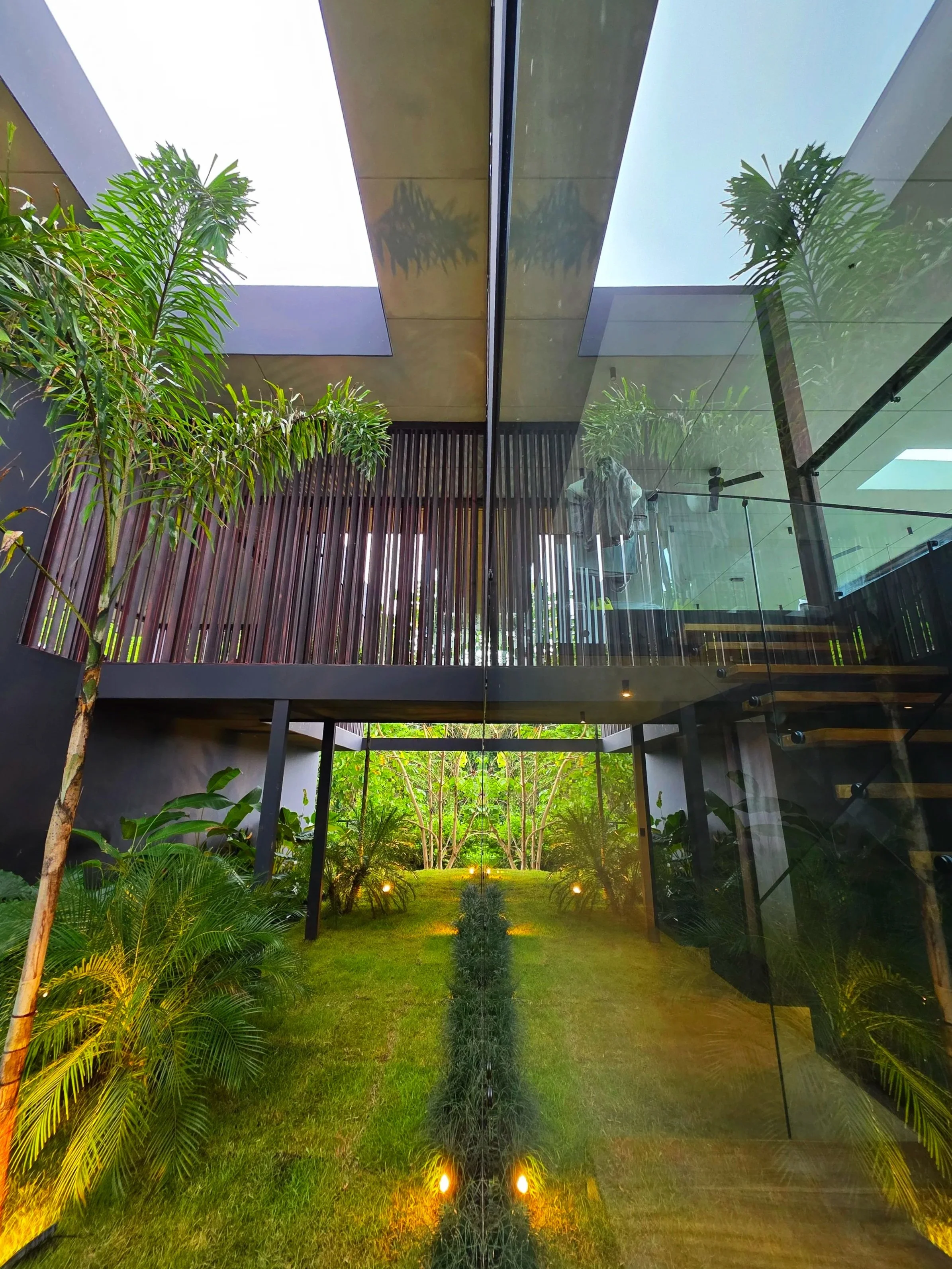 Interior view of a modern house with a glass wall, showcasing a green garden with trees and plants, and a staircase with wooden steps.