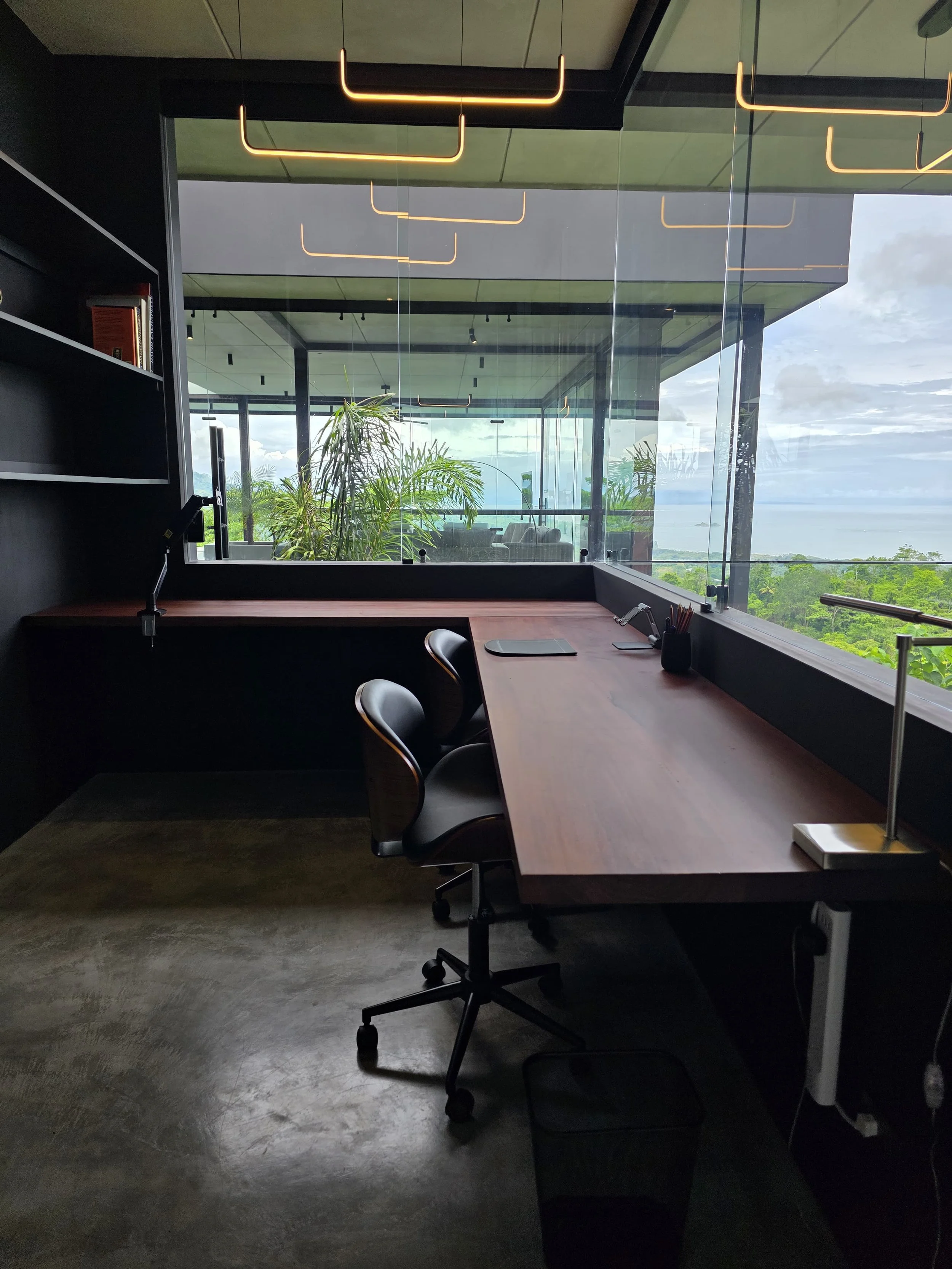 Modern office workspace with a wooden desk, black chairs, a balcony with glass railings, green plants, and a scenic view of the ocean and sky.
