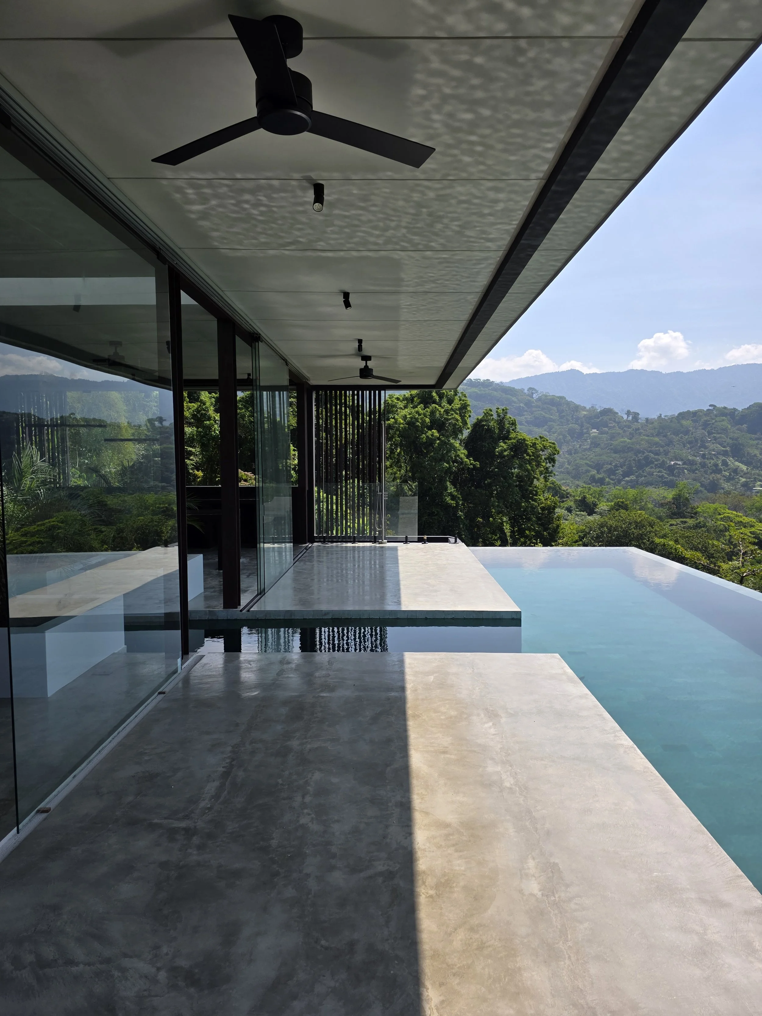 Modern outdoor balcony with sliding glass doors, ceiling fans, and a swimming pool overlooking lush green hills under a clear sky.
