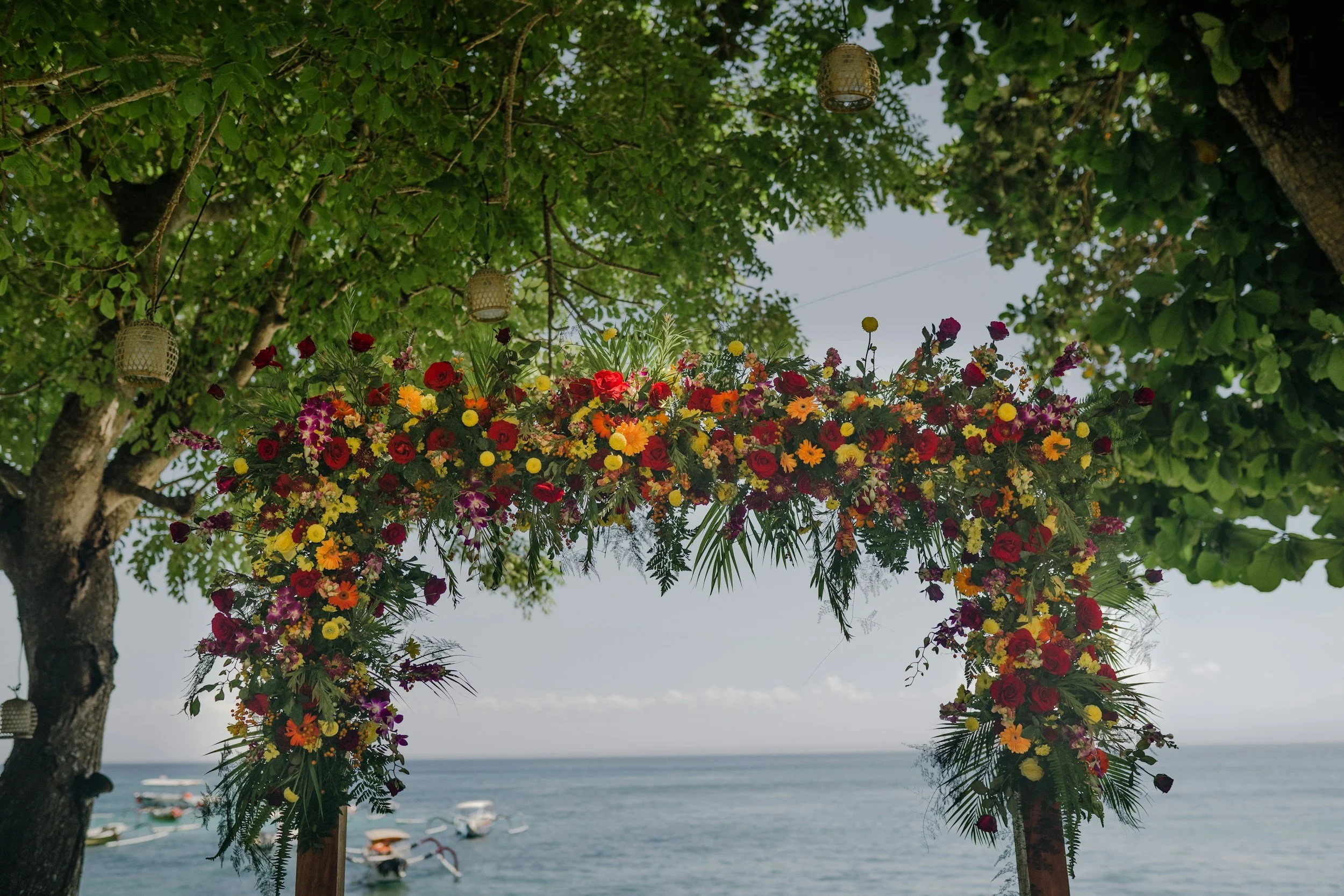A colorful floral wedding arch with red, yellow, and purple flowers, set against a lake view with boats, under trees with lanterns hanging from branches.