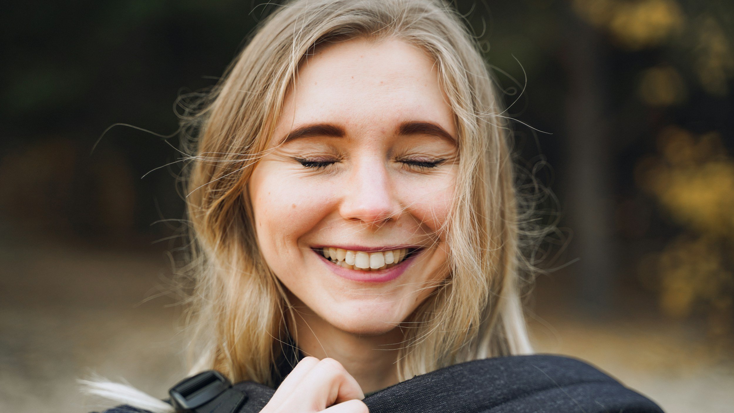A smiling woman with blonde hair and closed eyes outdoors during fall.