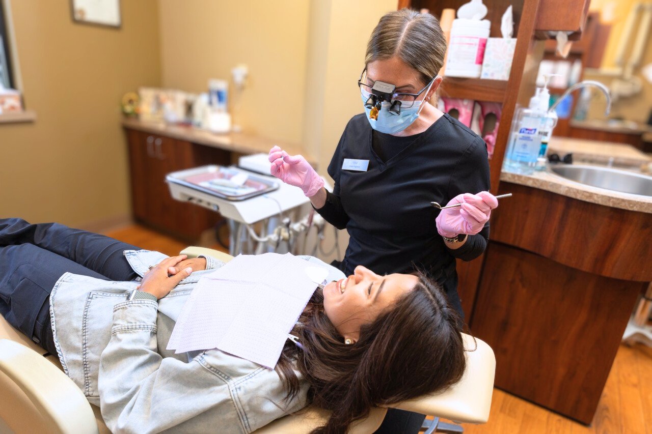 A woman lying on a dental chair at a dental office, receiving dental treatment from a dentist wearing magnifying glasses, a face mask, and gloves.
