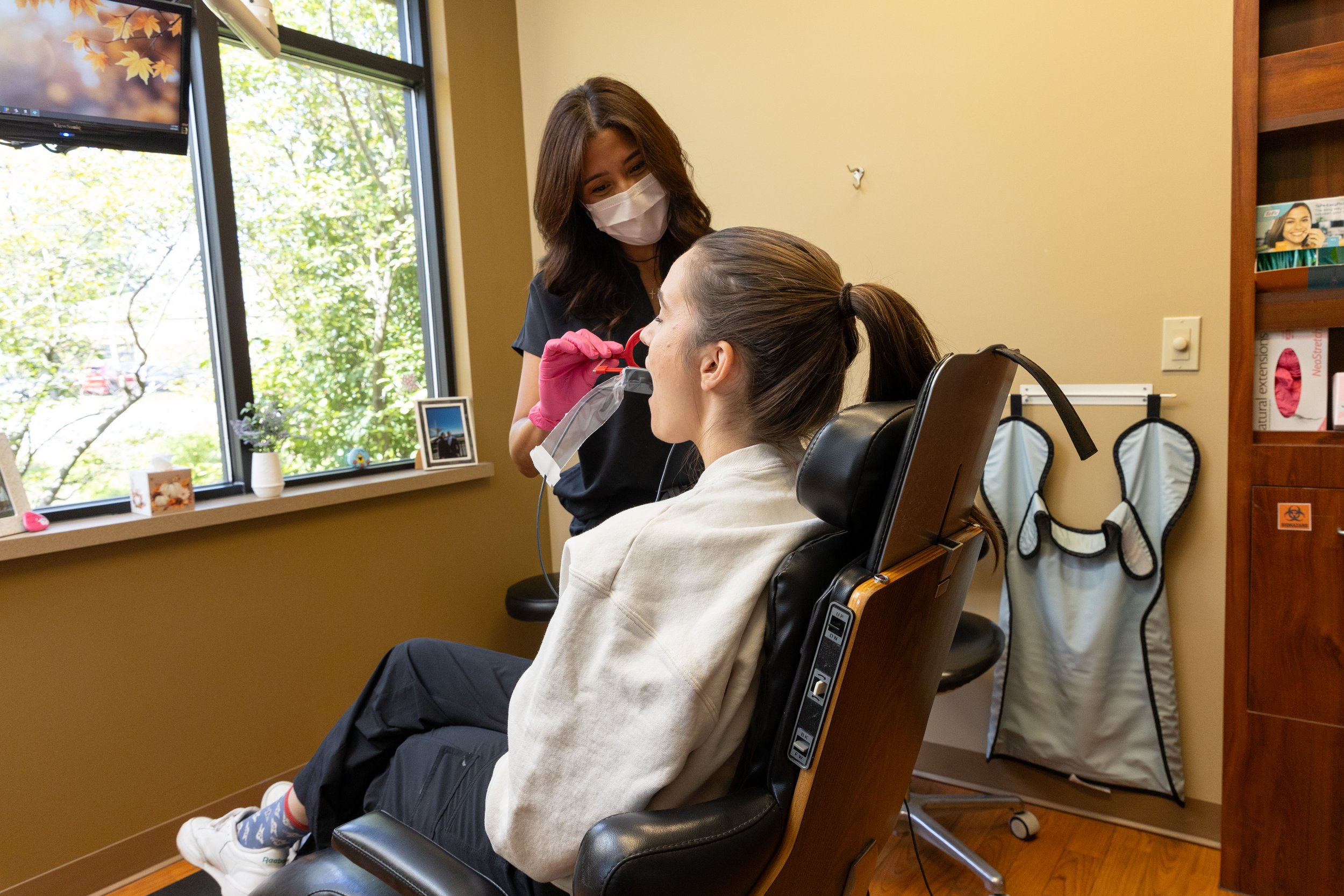 A woman is sitting in a dental chair with her mouth open, while a dental professional in a black uniform and pink gloves is preparing to examine her mouth.