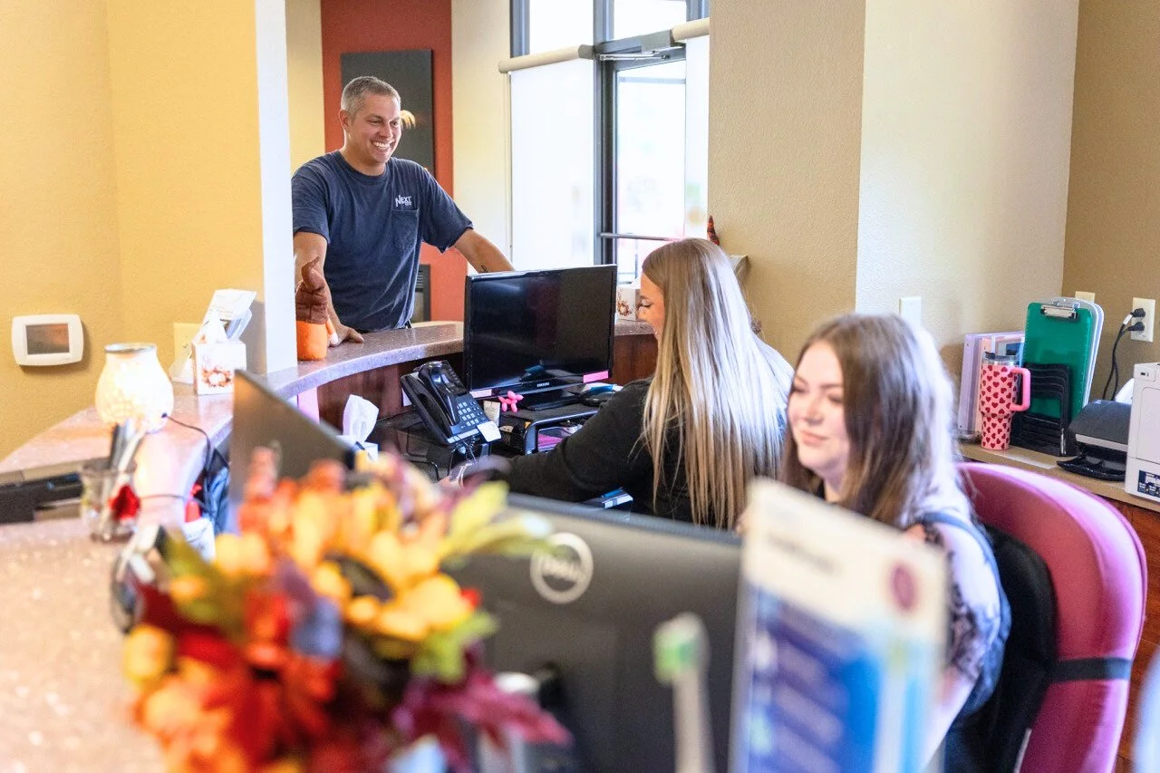 A man behind a reception desk talking to two women sitting at the desk, with office supplies and flowers in the foreground.