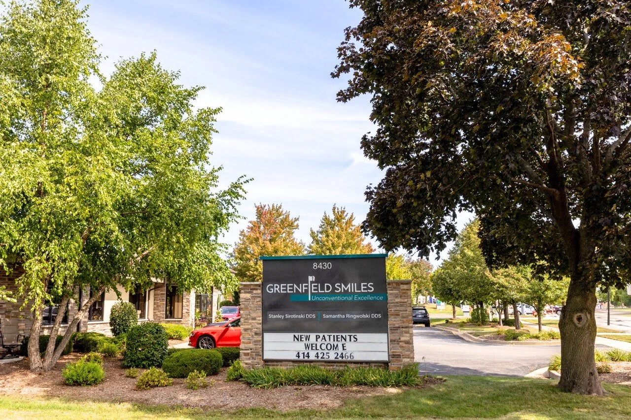 A sign for Greenfield Smiles dental office, located at 8430, surrounded by trees and shrubs, with parked cars nearby.