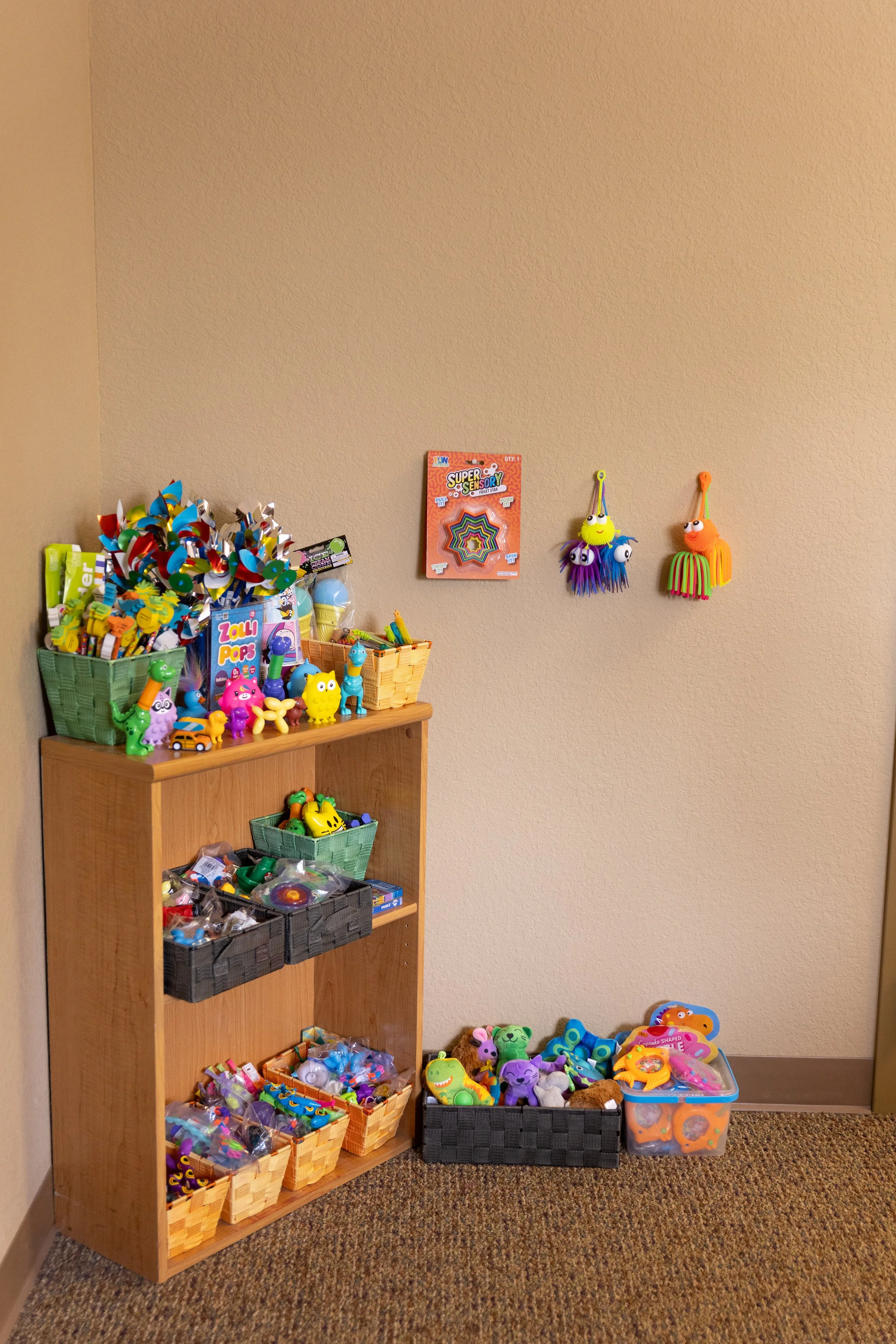 Colorful toys and prizes displayed on a wooden shelf and stored in baskets against a beige wall.