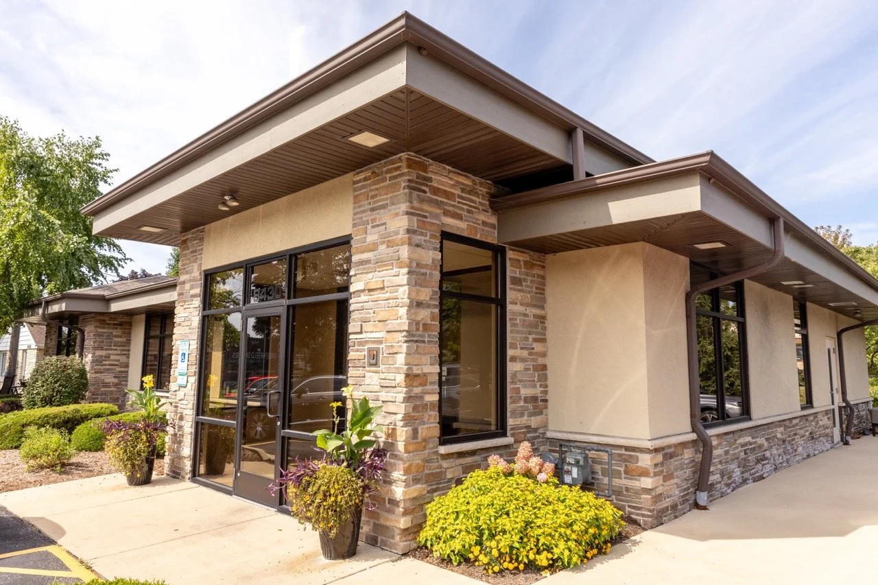 Exterior of a modern commercial building with brick and stucco walls, large glass windows, and landscaping featuring potted plants and flowering bushes.