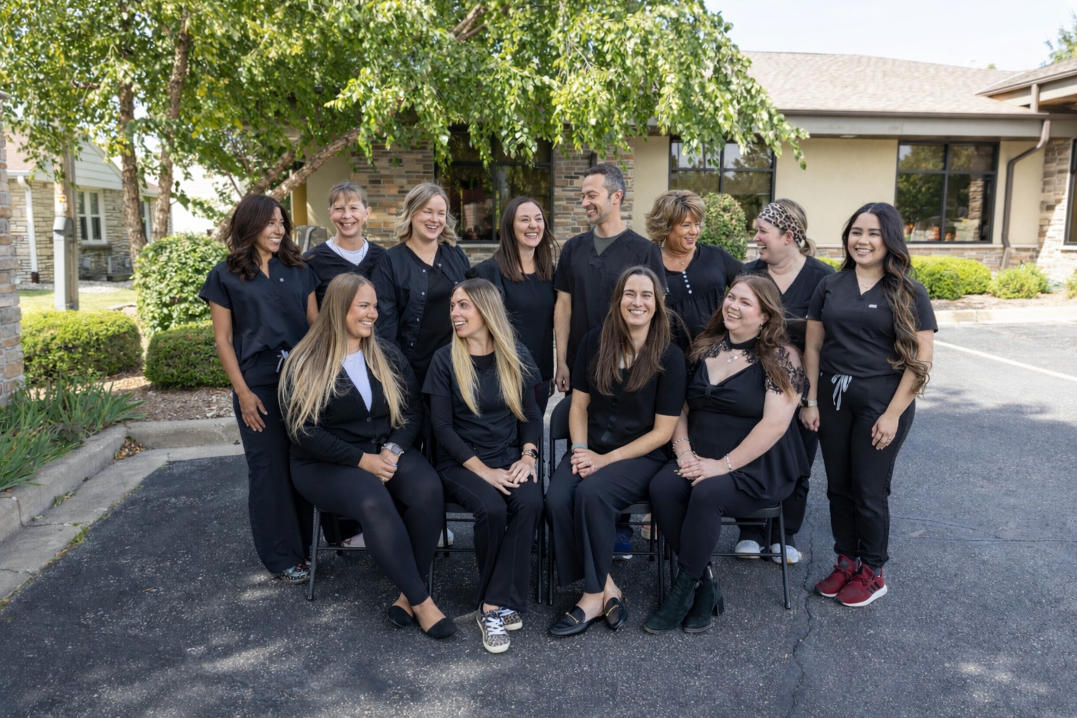 Group of medical professionals, mainly women, dressed in black scrubs, standing outside in front of a house, smiling and laughing.