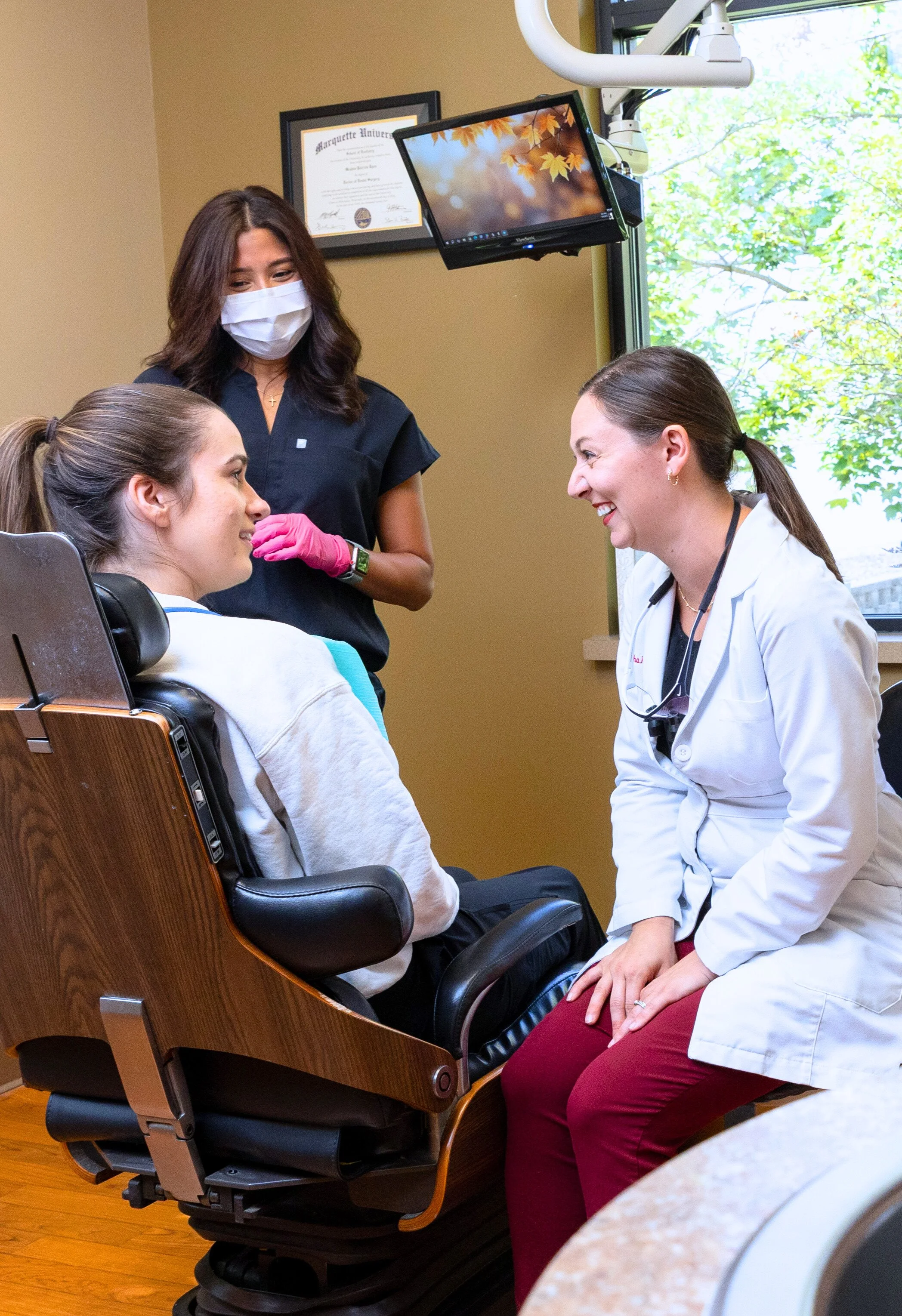 A woman in a white medical coat talking to a young woman sitting in a chair during a medical consultation. A healthcare professional standing nearby, wearing scrubs and a face mask, is observing. The room has a framed diploma on the wall and a window with a view of trees.