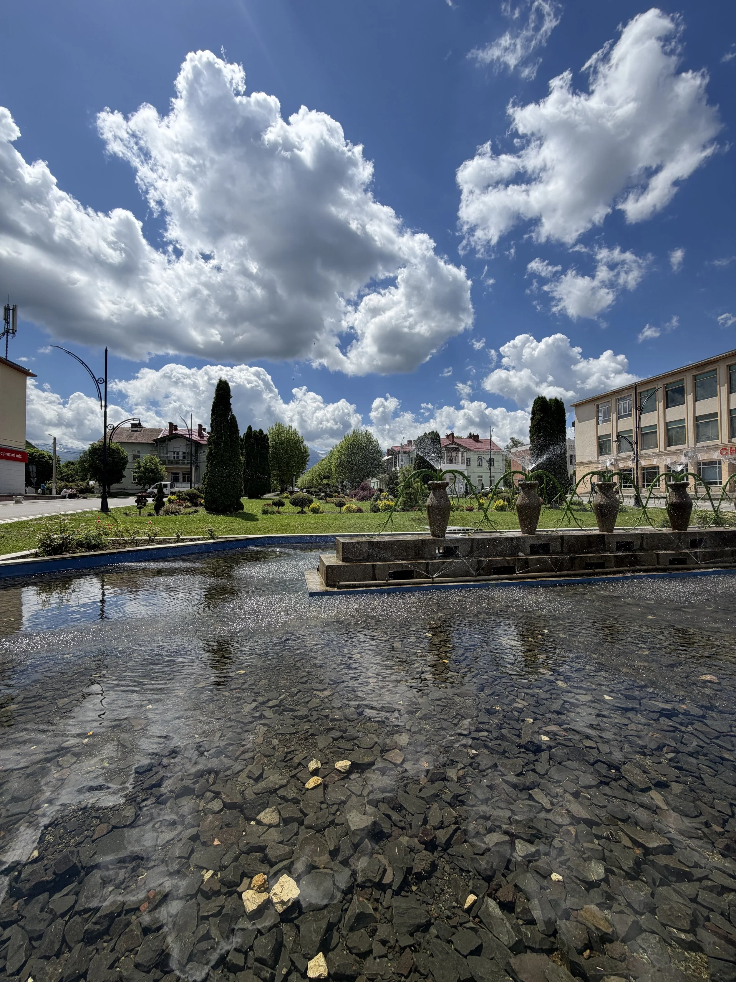 A small fountain with water jets in a park, surrounded by green grass, trees, residential buildings, and a bright sky with fluffy clouds.