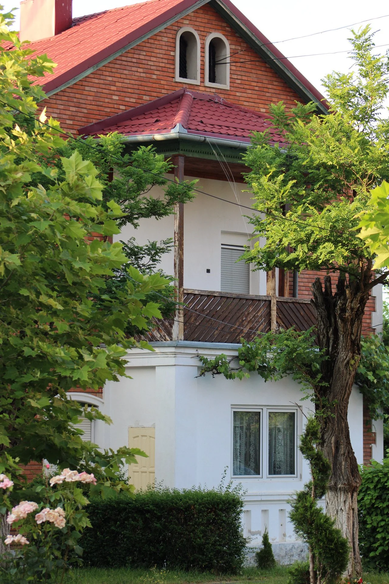 A house with brick and white exterior walls, framed by green trees and shrubs, and a red tiled roof.