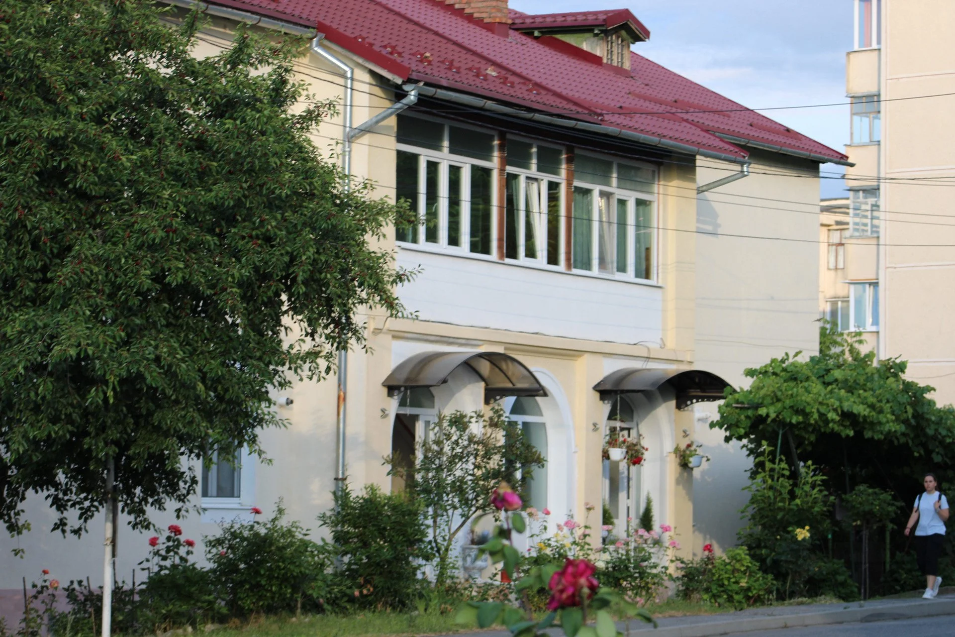 A residential house with a red metal roof and several large windows, surrounded by a lush garden with trees and colorful flowers. A person wearing a white shirt and black pants is walking on the sidewalk beside the house.