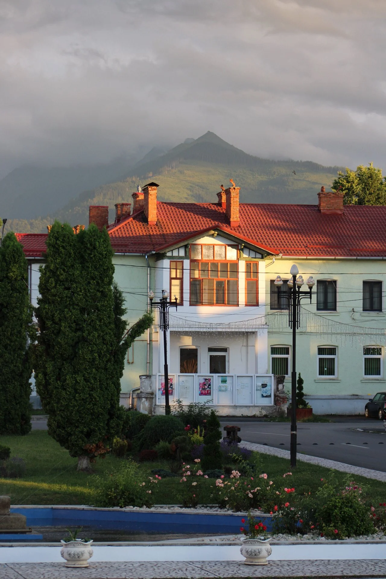 A house with red tiled roof and brick chimneys, set against a mountain background with a cloudy sky, surrounded by trees and a manicured garden with flowers and a fountain.