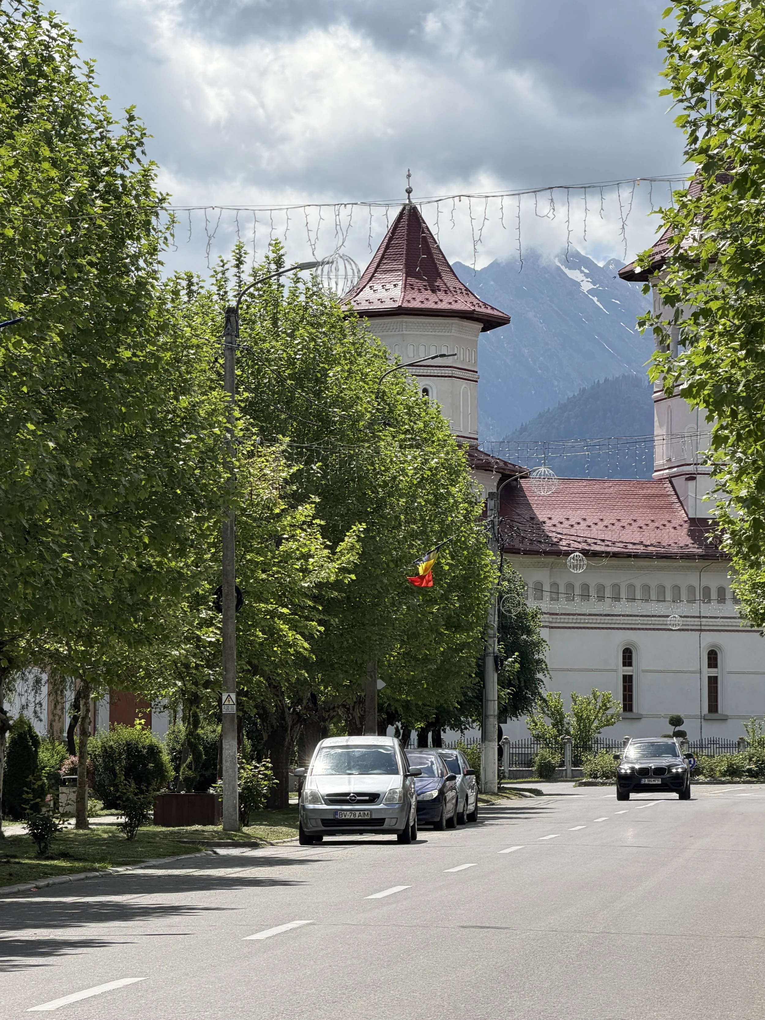 A street view with parked cars on the side, trees lining the sidewalk, a church with a red roof and white walls, decorative string lights, and mountains in the background under a cloudy sky.