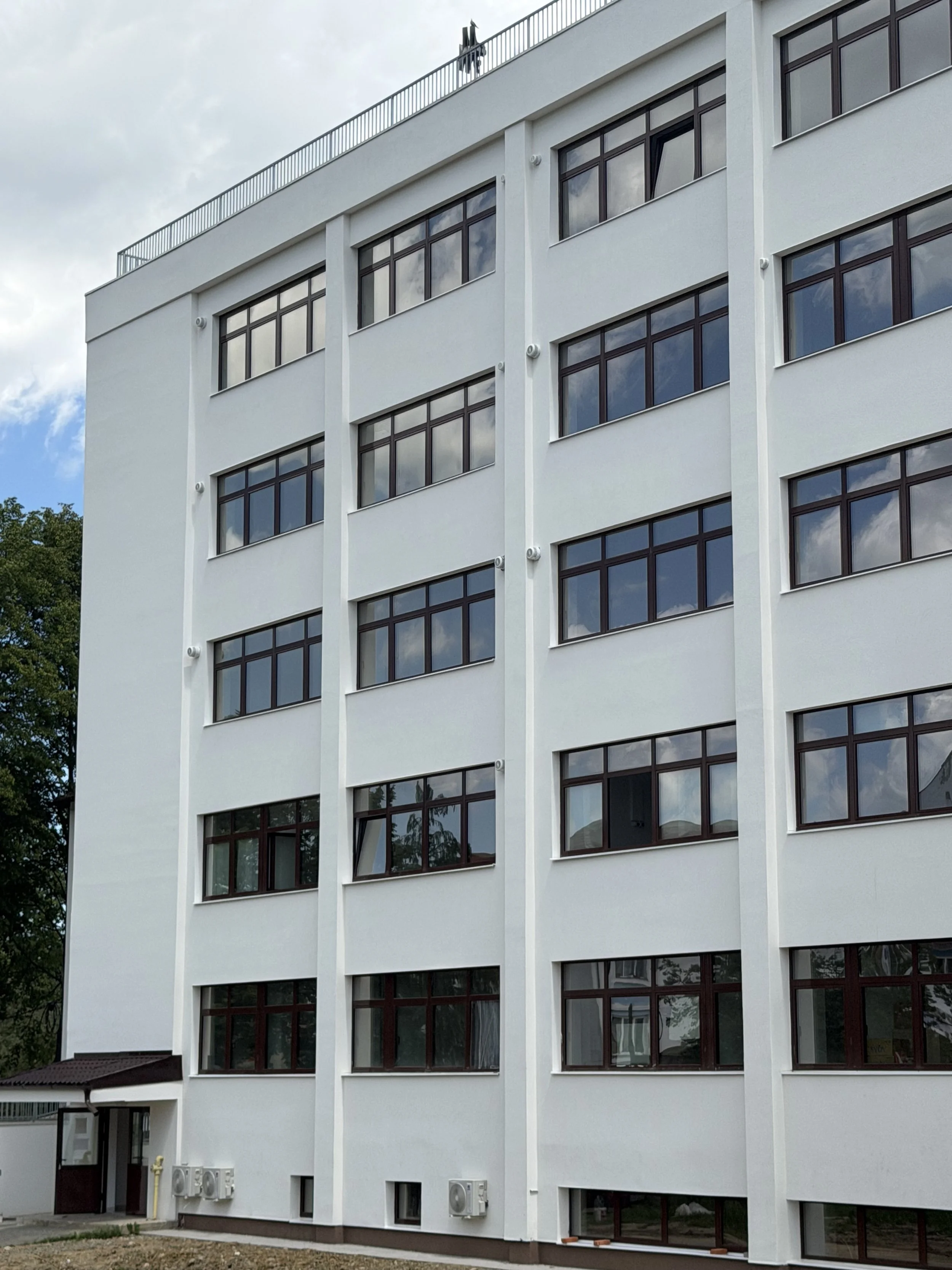 Modern white multi-story building with large windows, some with reflection of clouds, green trees in the background.
