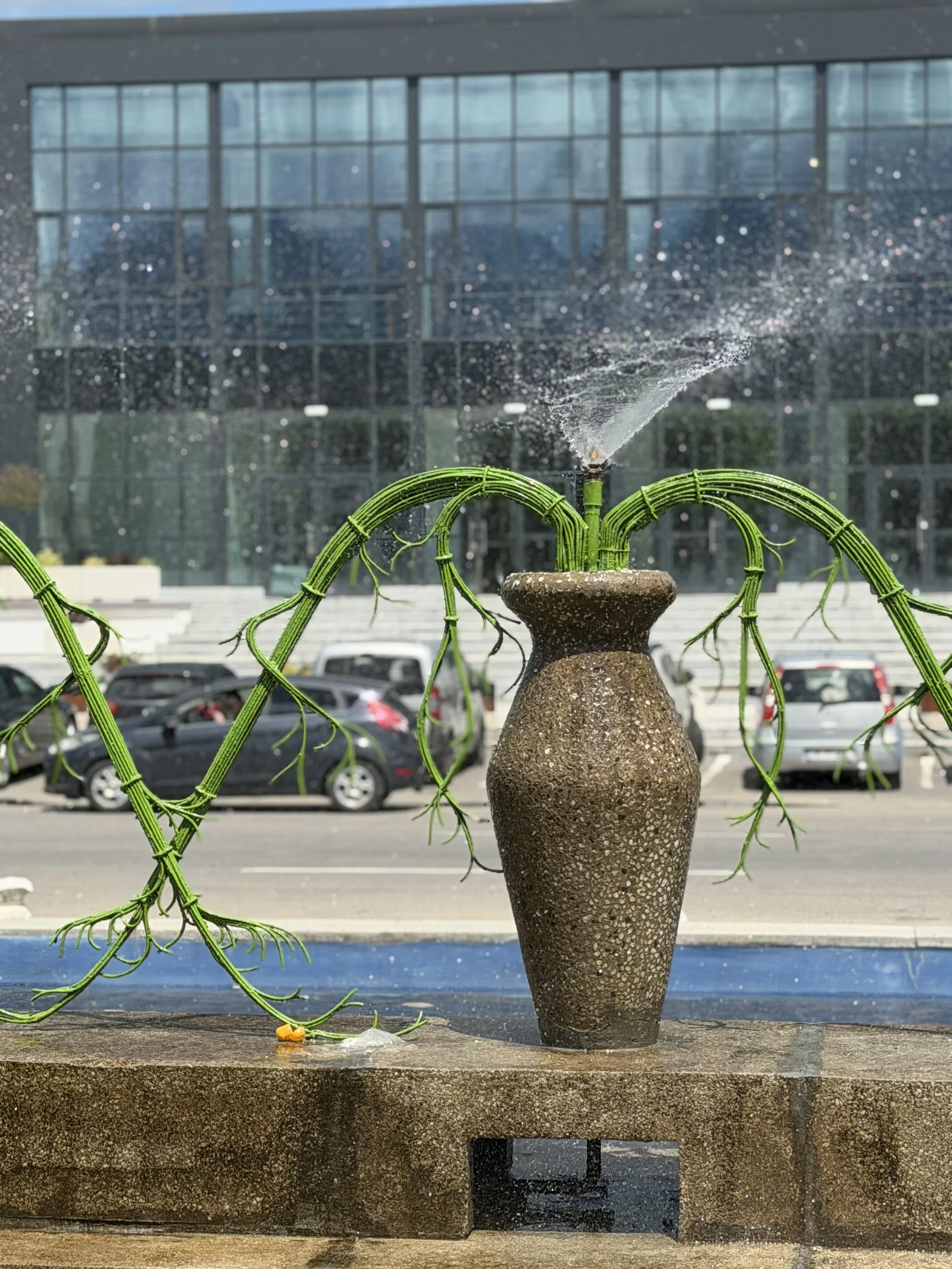 A decorative fountain with a large ceramic vase-like base and a small sculpture on top, from which water sprays outward. Green ornamental metalwork surrounds the fountain, set against a background of a modern glass building and a parking lot with car