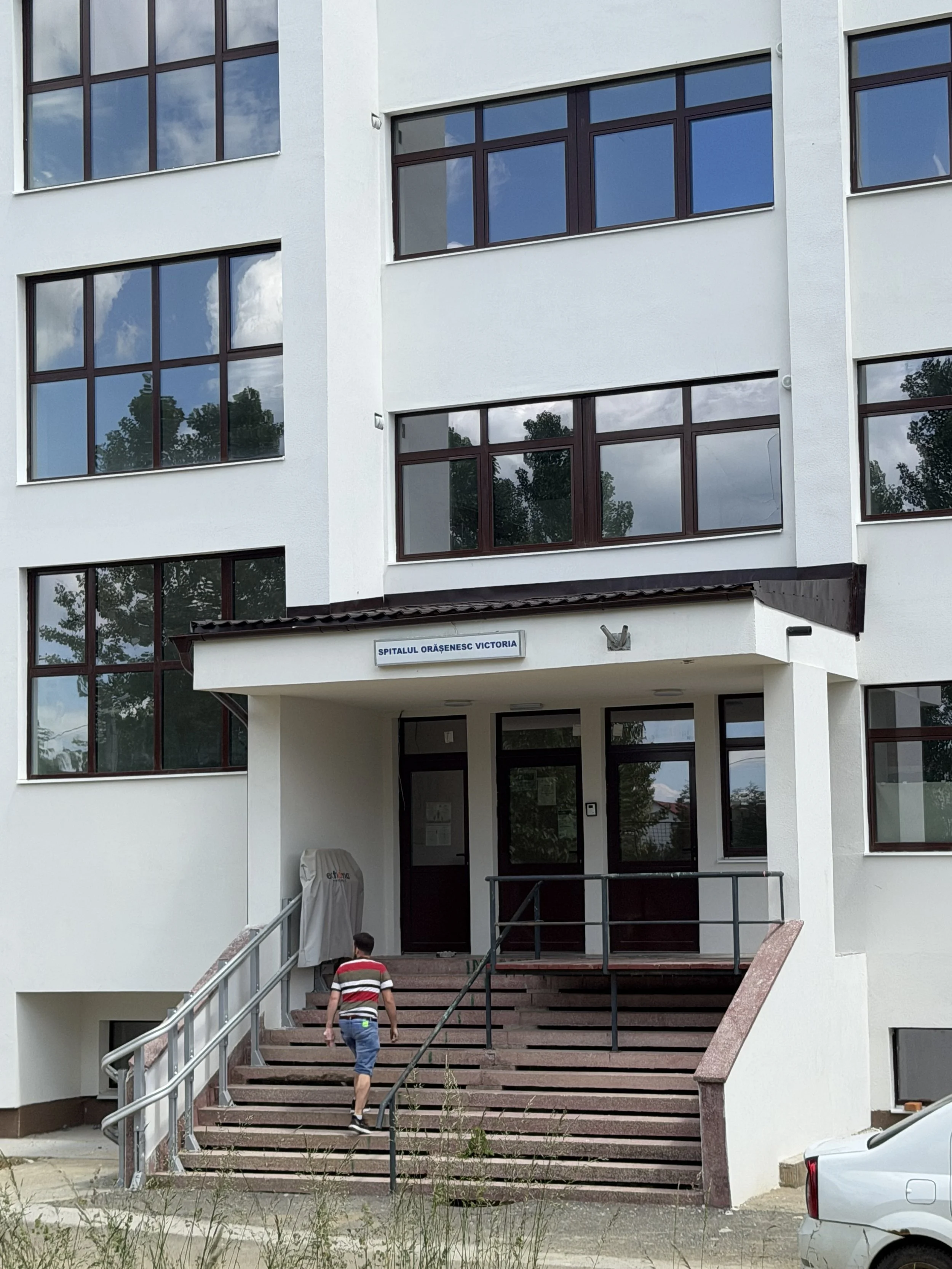 Front view of a modern white building with large windows reflecting clouds and trees, a sign reading 'Spitalul Orășenesc Victoria' above the entrance, stairs leading up to the door, and a man walking towards the entrance.