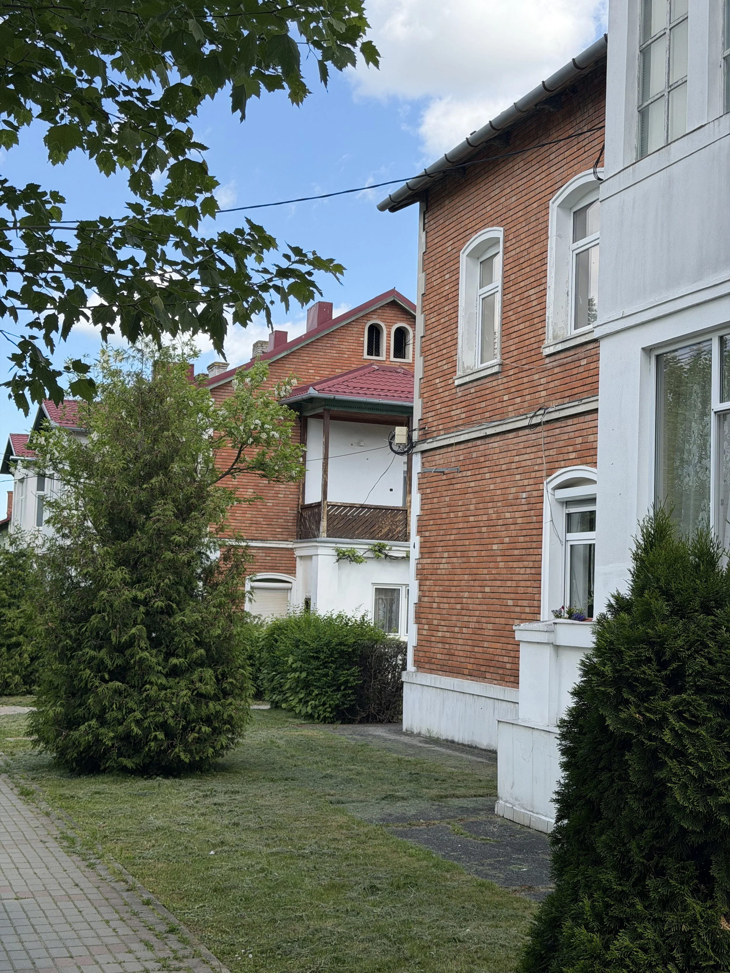 View of residential buildings with red brick and white exteriors, surrounded by green trees and grass, under a partly cloudy sky.