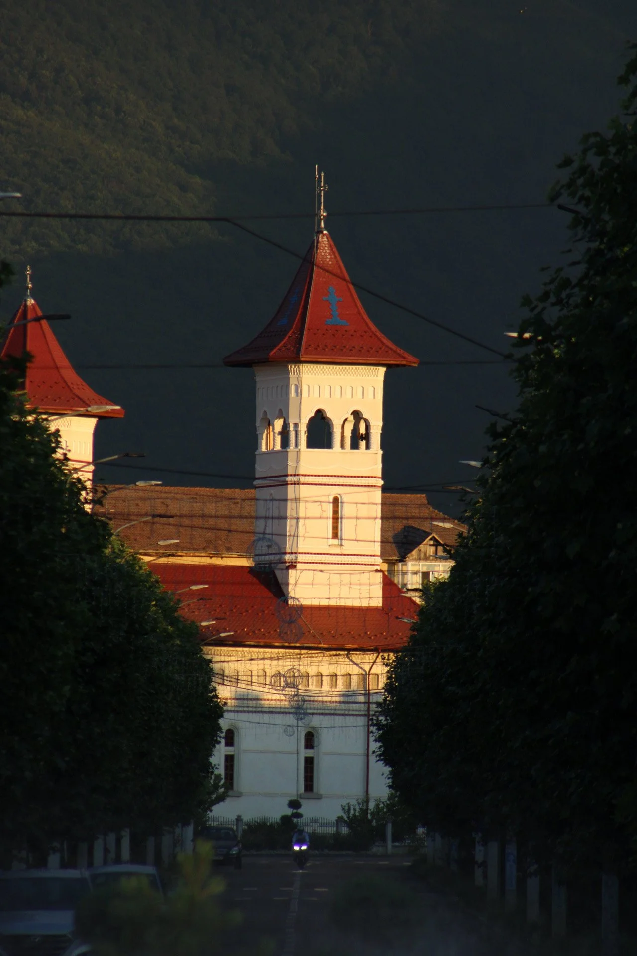 A tall white church with a red roof and bell tower, framed by trees on a street at sunset with mountains in the background.