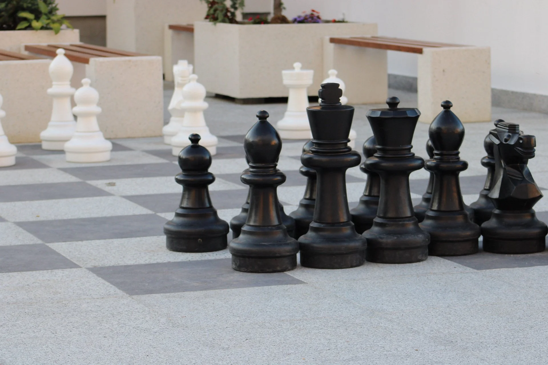 Large outdoor chess set with black and white chess pieces on a checkered concrete surface, surrounded by concrete planters with flowers and greenery.