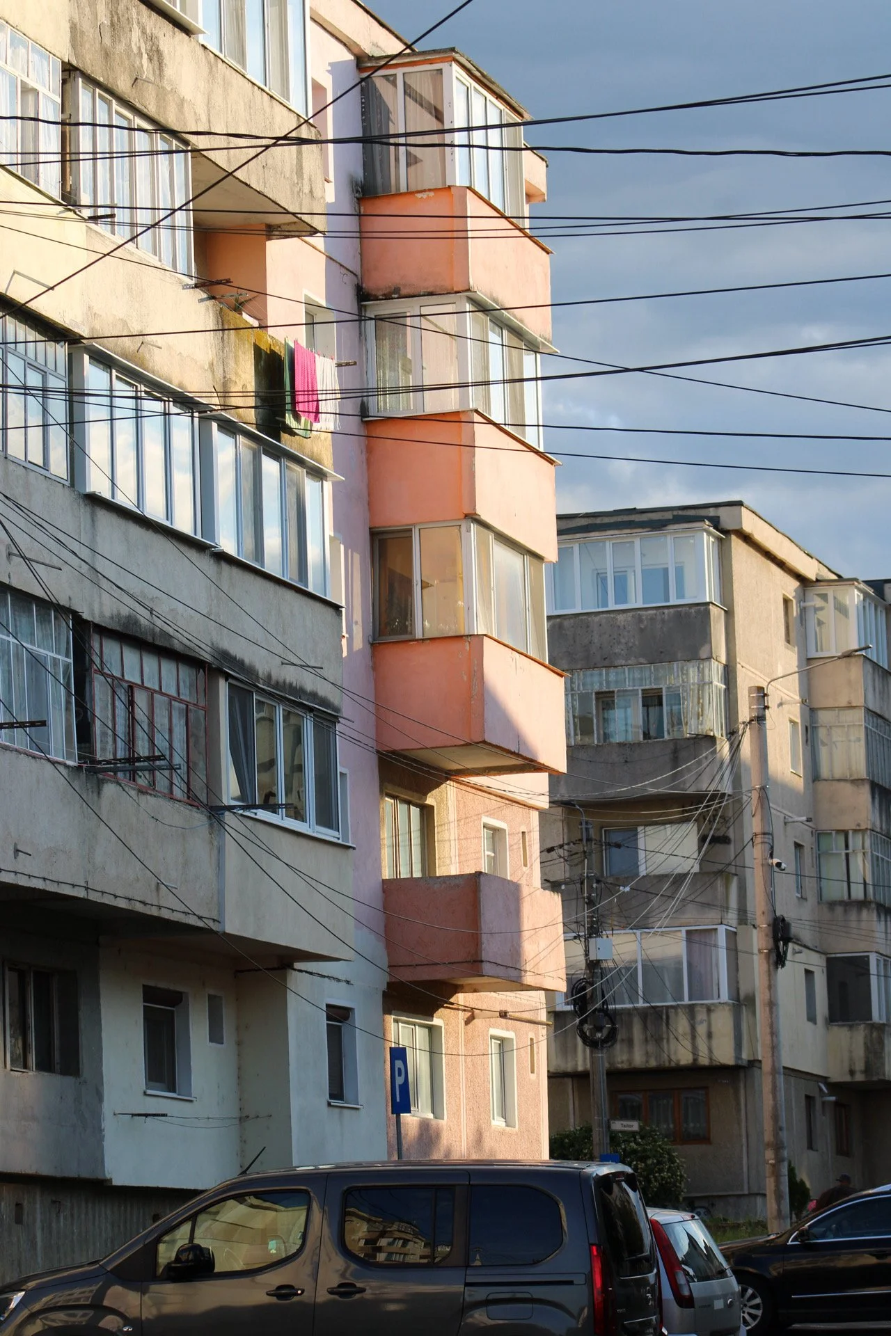 Apartment buildings with multiple floors and balconies, some enclosed with glass, with laundry hanging outside and electrical wires crossing in the foreground, parked cars at the bottom, under a cloudy sky.