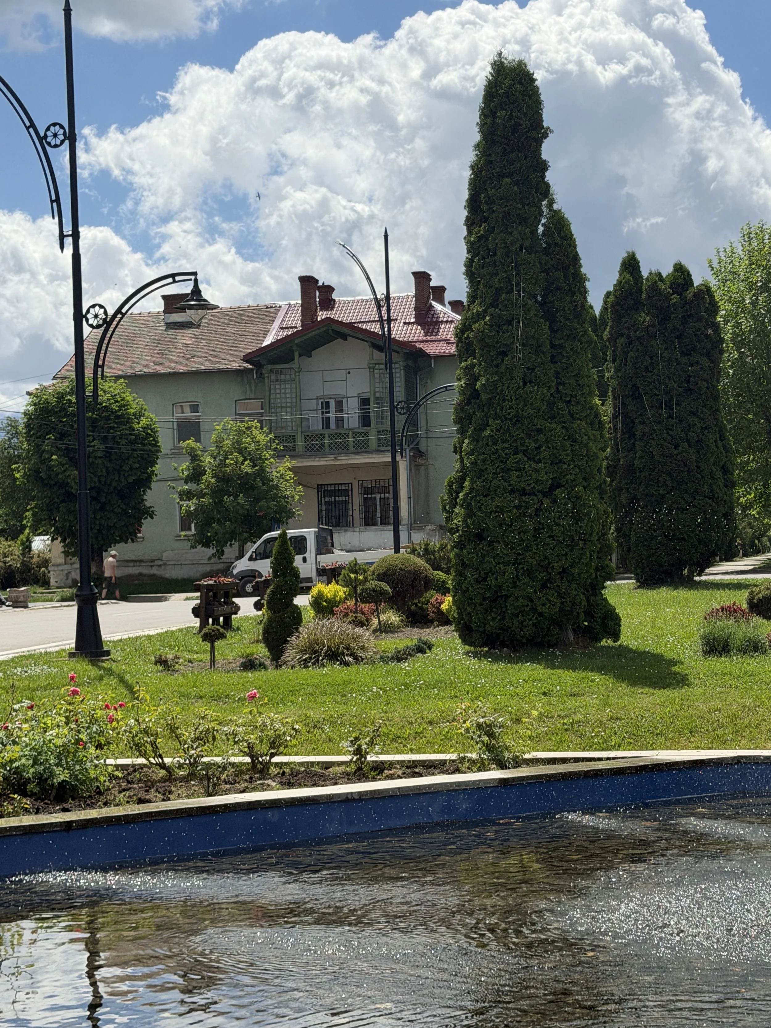 View of a house with a red roof behind lush green trees and a small pond in the foreground, under a sky with fluffy clouds.
