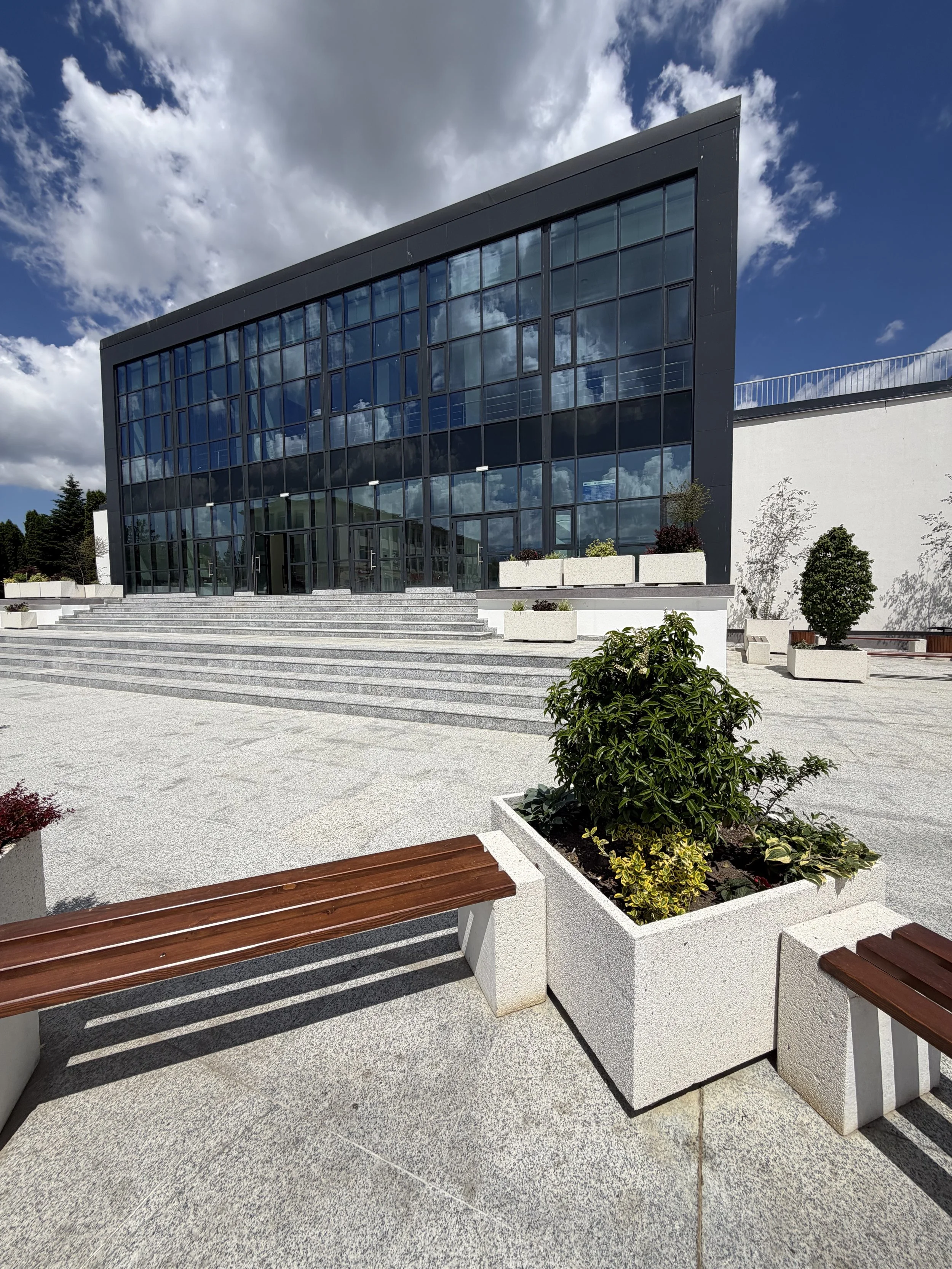 Modern building with large glass windows, steps leading up to entrance, and outdoor seating area with benches and potted plants, under partly cloudy sky.