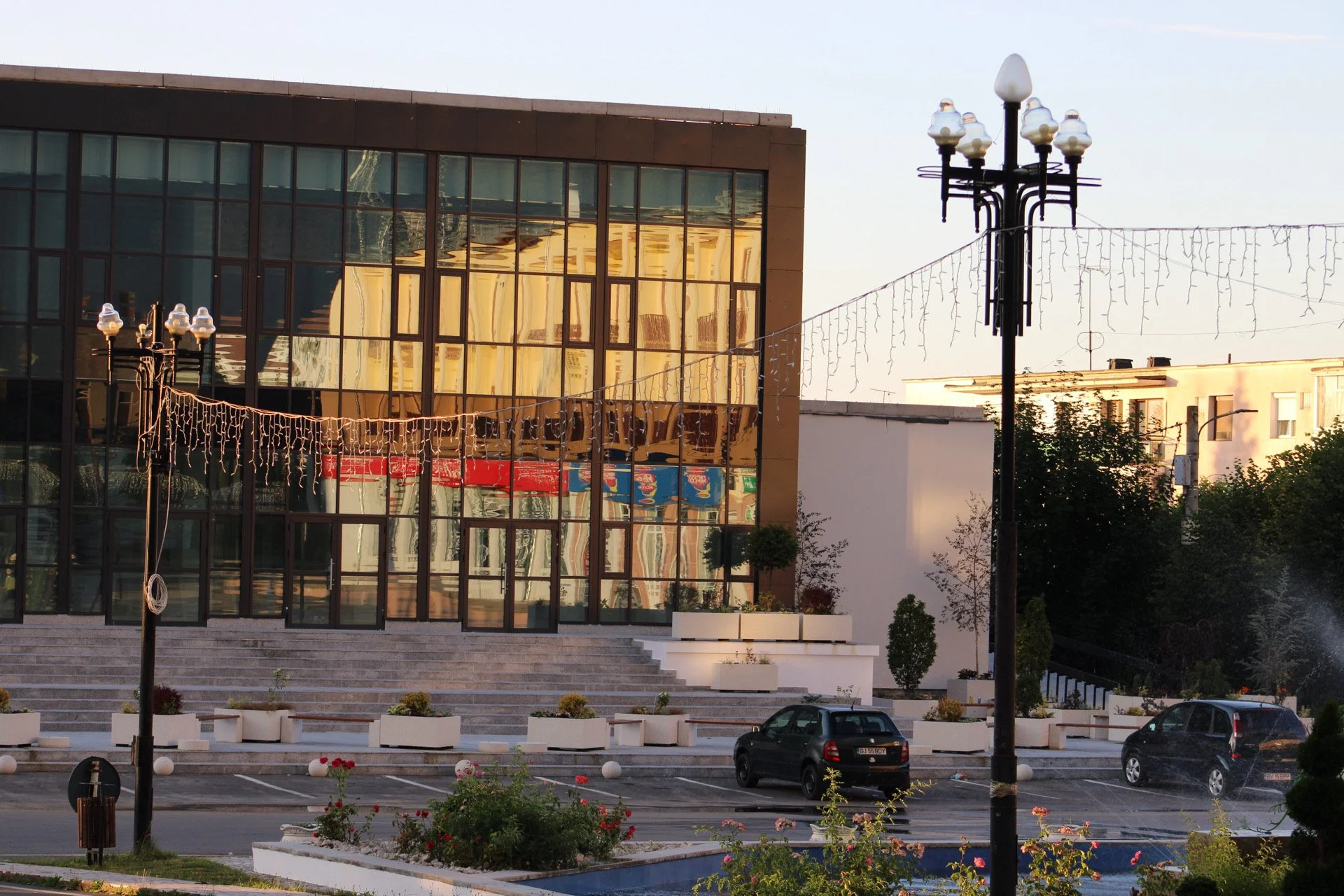 A modern glass building reflecting other buildings in its windows, with a step entrance, decorated with string lights and potted plants outside, and parked cars in front, illuminated by warm sunlight in the late afternoon or early evening.