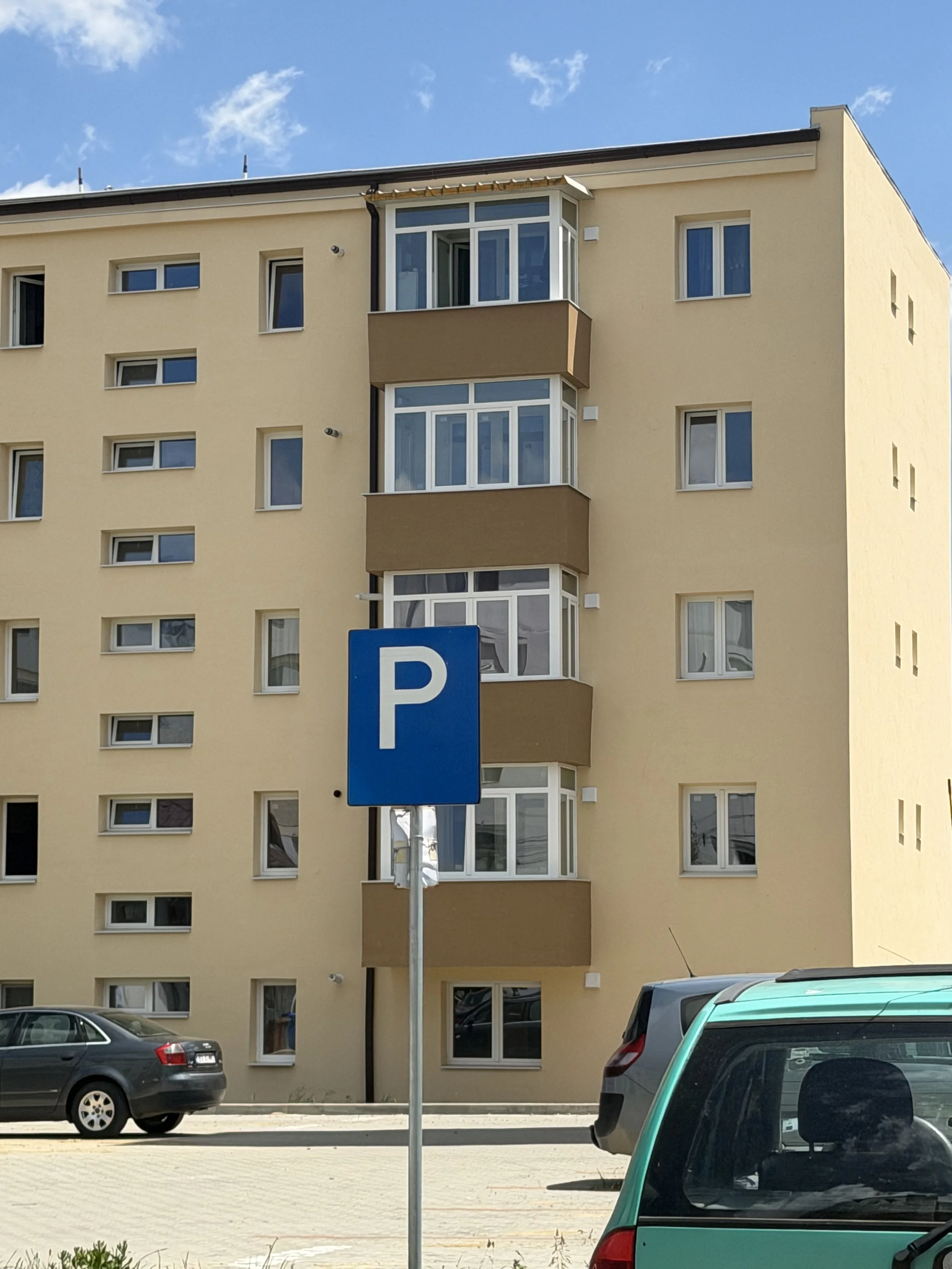 A residential apartment building with several windows and balconies. There is a parking lot in front with cars, and a blue parking sign with a white 'P'. The sky is blue with some clouds.