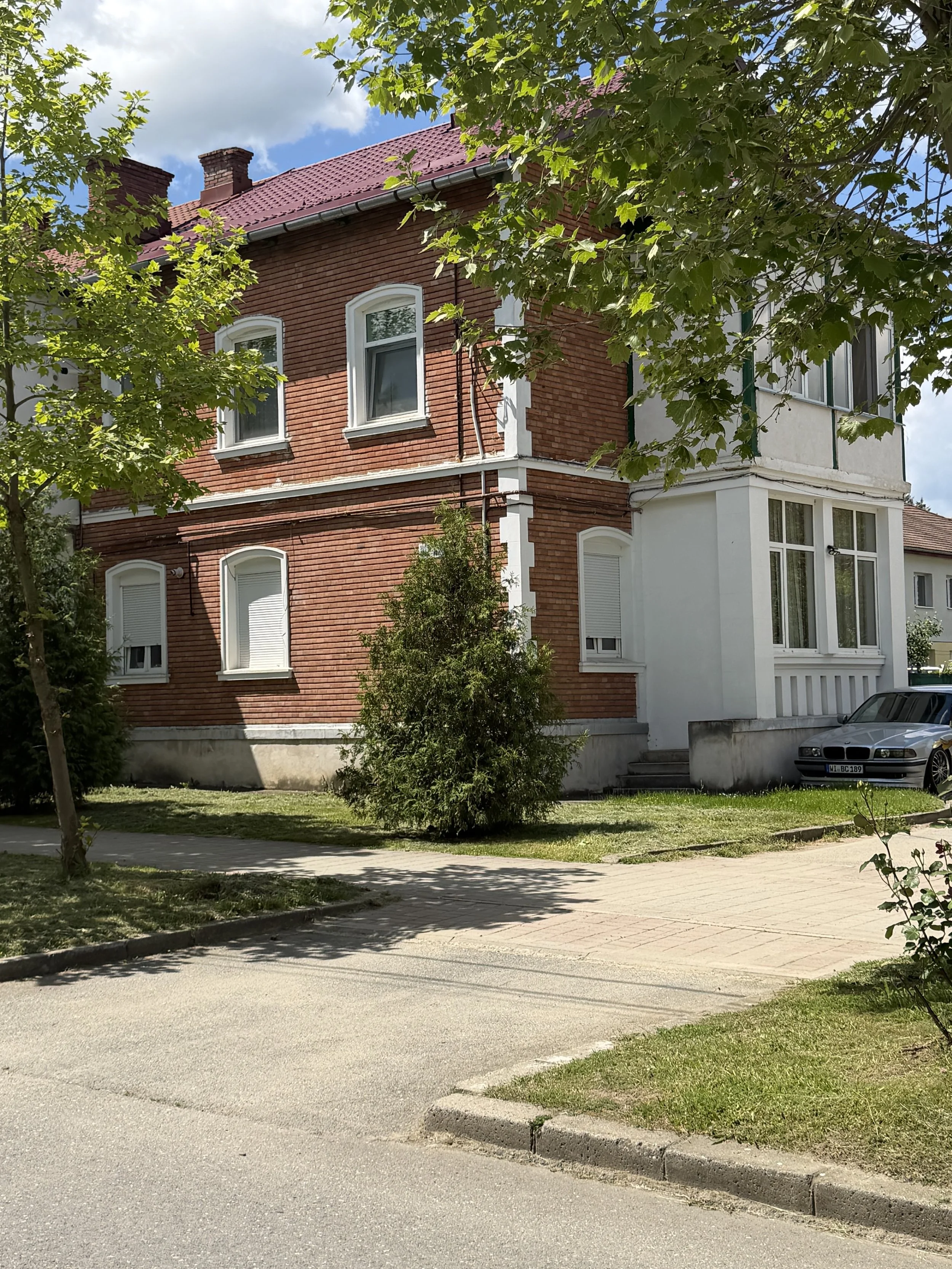 A residential area with a brick house, a car parked on the street, green trees, and a partly cloudy sky.
