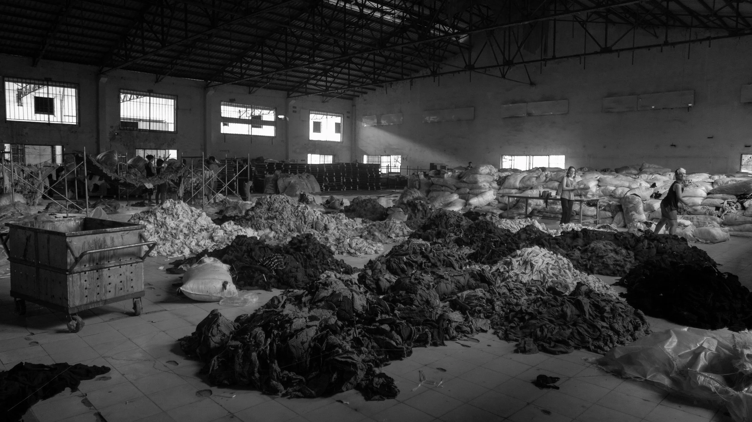 Interior of a large warehouse with piles of various clothes, workers sorting through fabric, and stacks of bags on the floor.